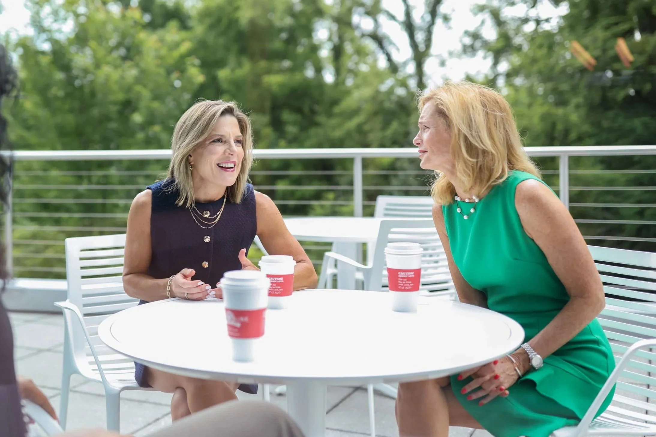 Two women are sitting at a white outdoor table, engaged in conversation. One woman, with shoulder-length blonde hair and wearing a dark sleeveless top, is smiling and holding a disposable coffee cup. The other woman, with curly blonde hair and wearing a green sleeveless dress with a necklace, is leaning forward and listening. There are similar disposable coffee cups on the table, and the background shows green trees and outdoor furniture.
