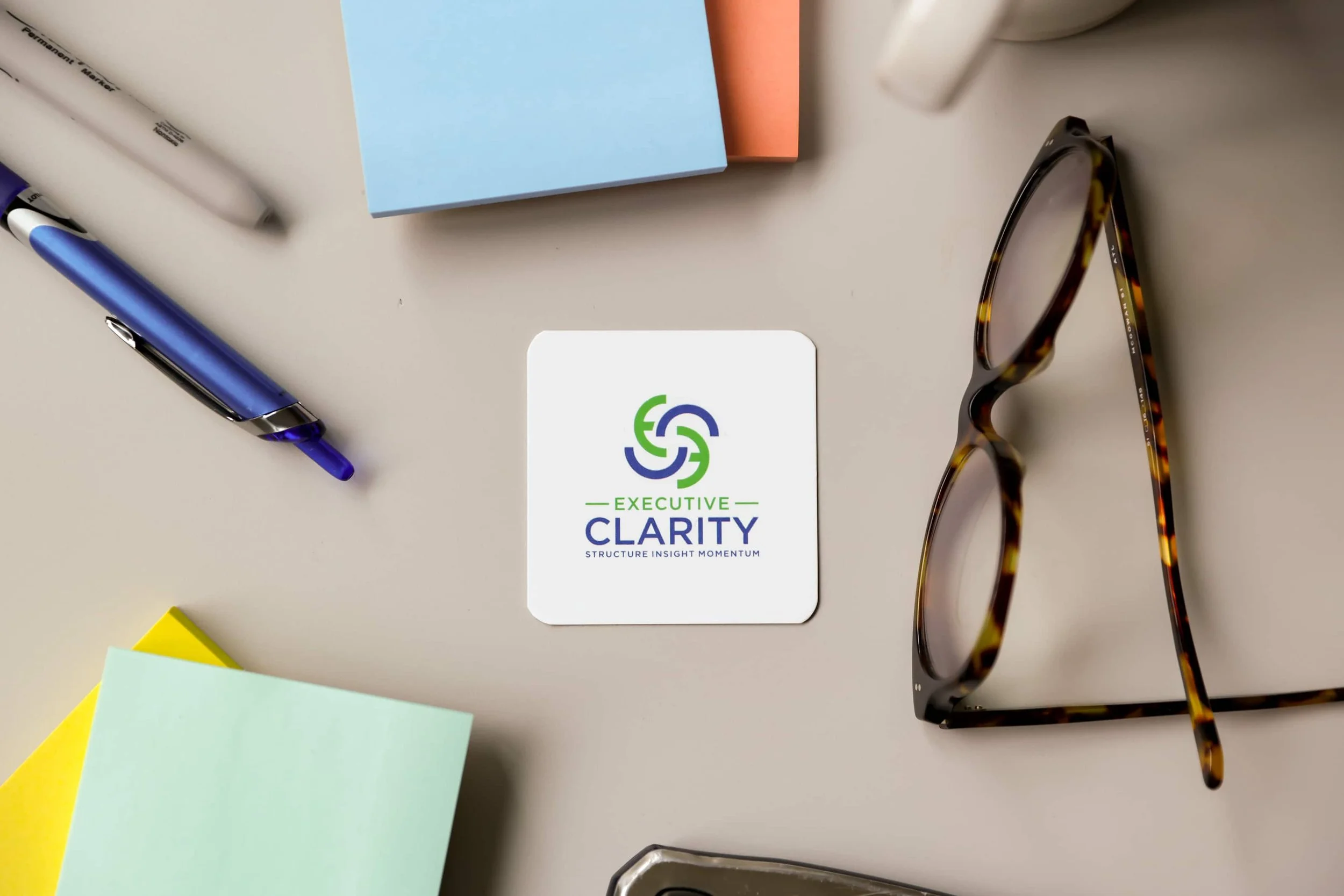 An overhead view of a desk with office supplies, including a blue and a pink sticky note pad, a blue pen, a pair of tortoiseshell glasses, a white card with the logo and text 'Executive Clarity,' and several small sheets of colored paper in yellow, green, and light blue.