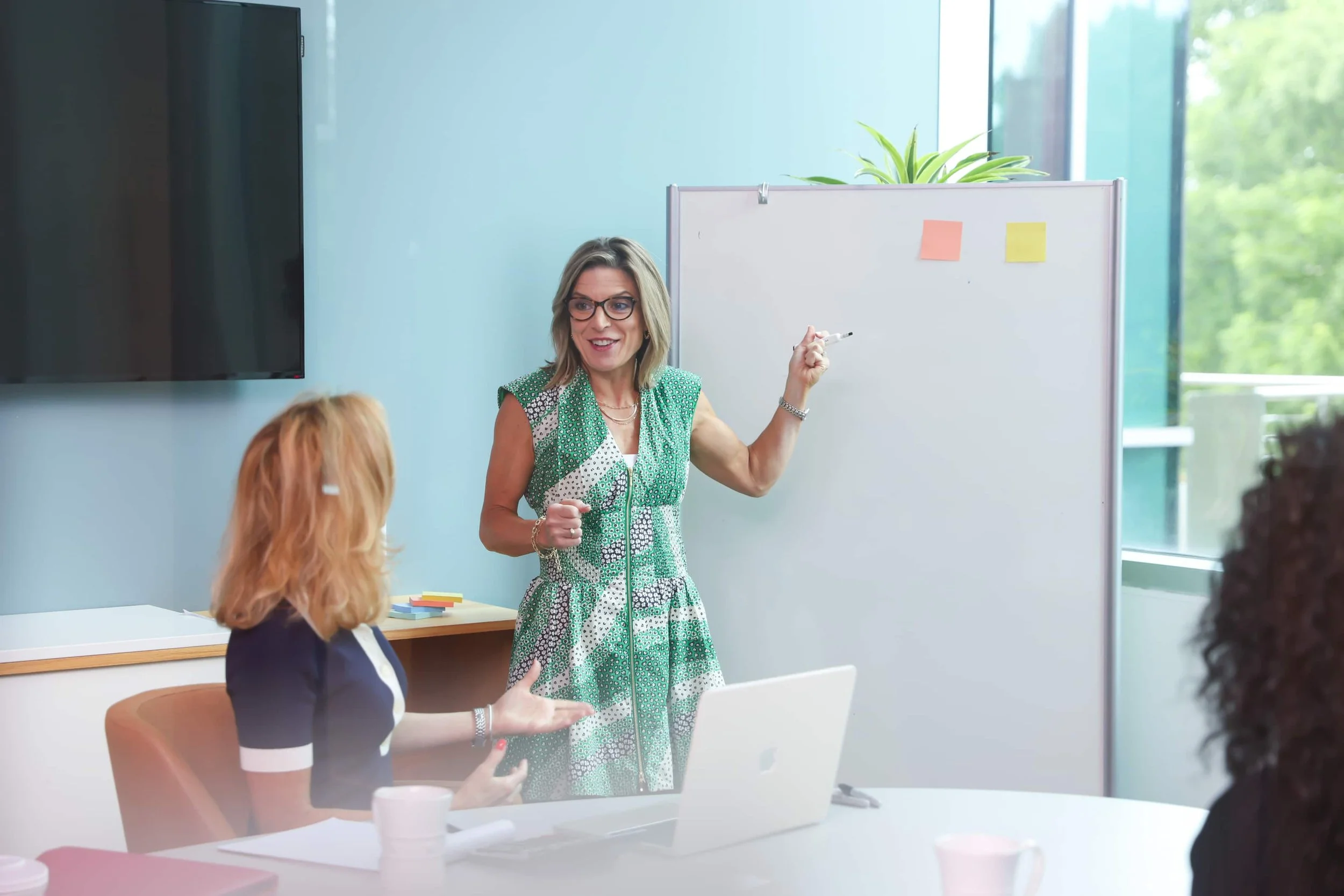 A woman with blonde hair, wearing glasses and a green patterned dress, is standing and smiling while gesturing with a marker in her hand in a modern conference room. Several people are sitting at a table with laptops and coffee cups, listening to her. There is a whiteboard with sticky notes and a large window with trees outside in the background.