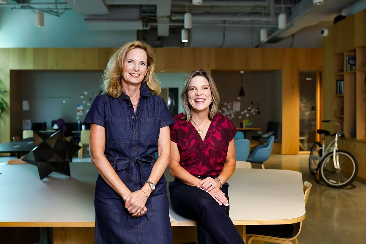Two women smiling in a modern office space, one standing and one sitting on a table, with a bicycle in the background.