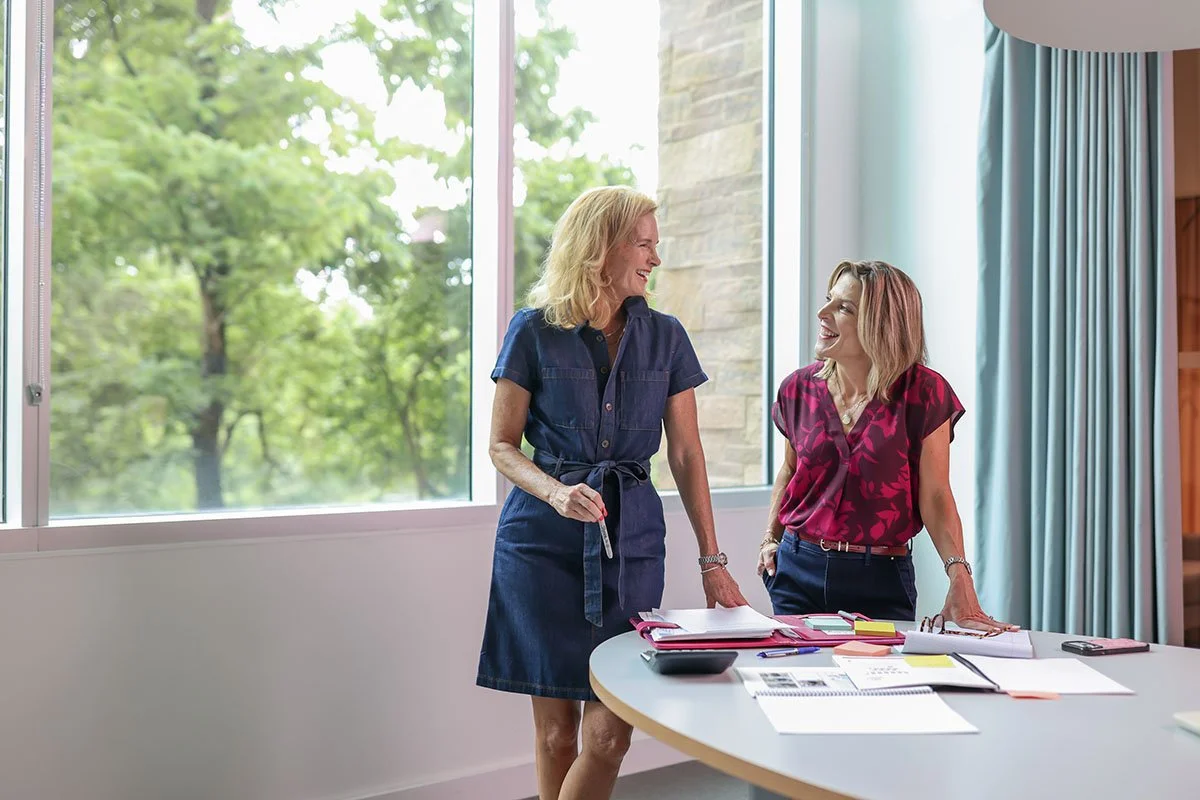Two women standing and smiling at each other in a bright conference room with a large window and green trees outside, surrounded by documents, notebooks, and stationery on a table.