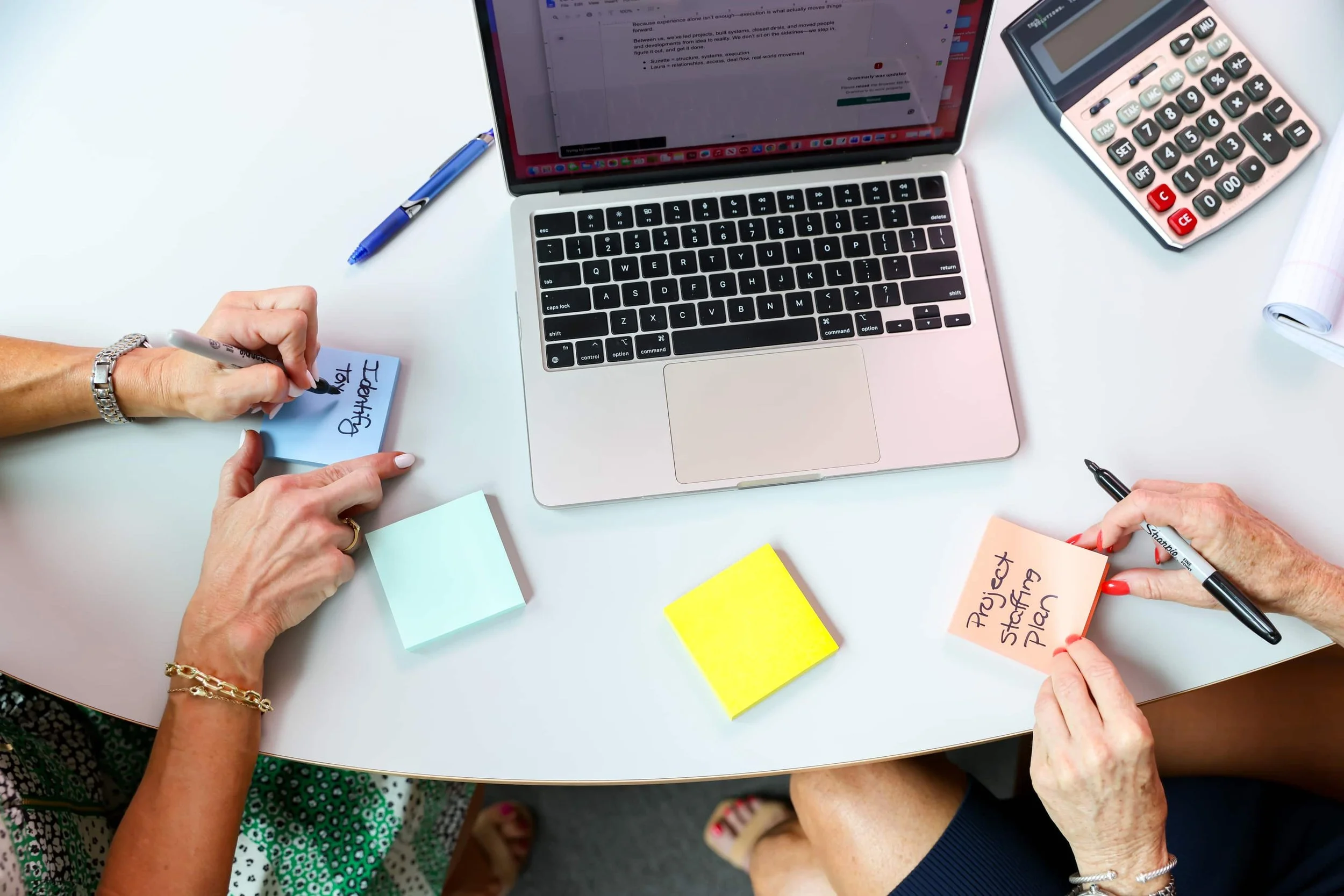 People working together at a white table with a laptop, calculator, sticky notes, pens, and a marker, writing on sticky notes.