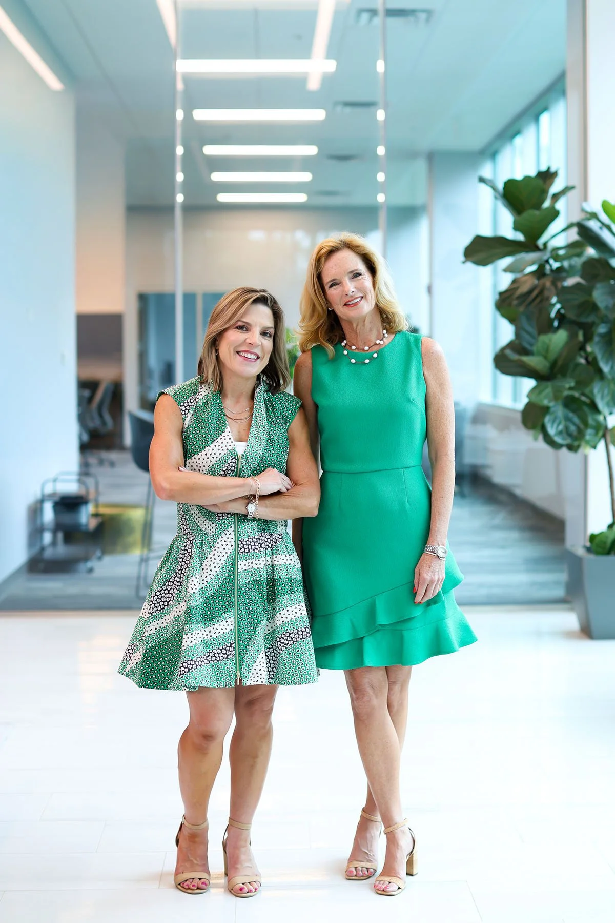 Two women smiling and standing together in an office hallway, both wearing green dresses and beige high heels. There are large windows and plants in the background.