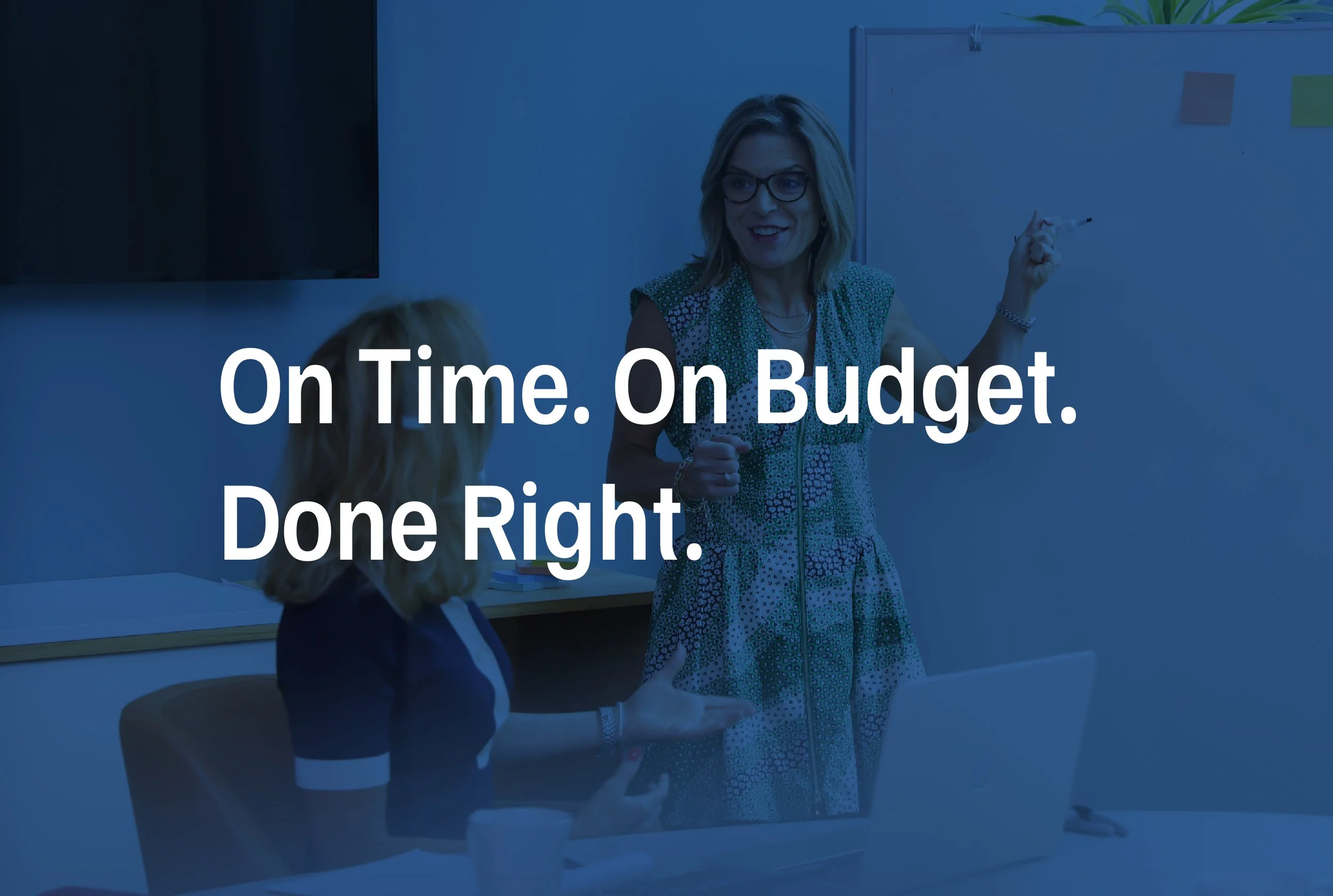 A woman with blonde hair, glasses, and a patterned dress is standing in a classroom, smiling and pointing at a whiteboard. A person with blonde hair is sitting in front of her. Text overlay says, 'On Time. On Budget. Done Right.'