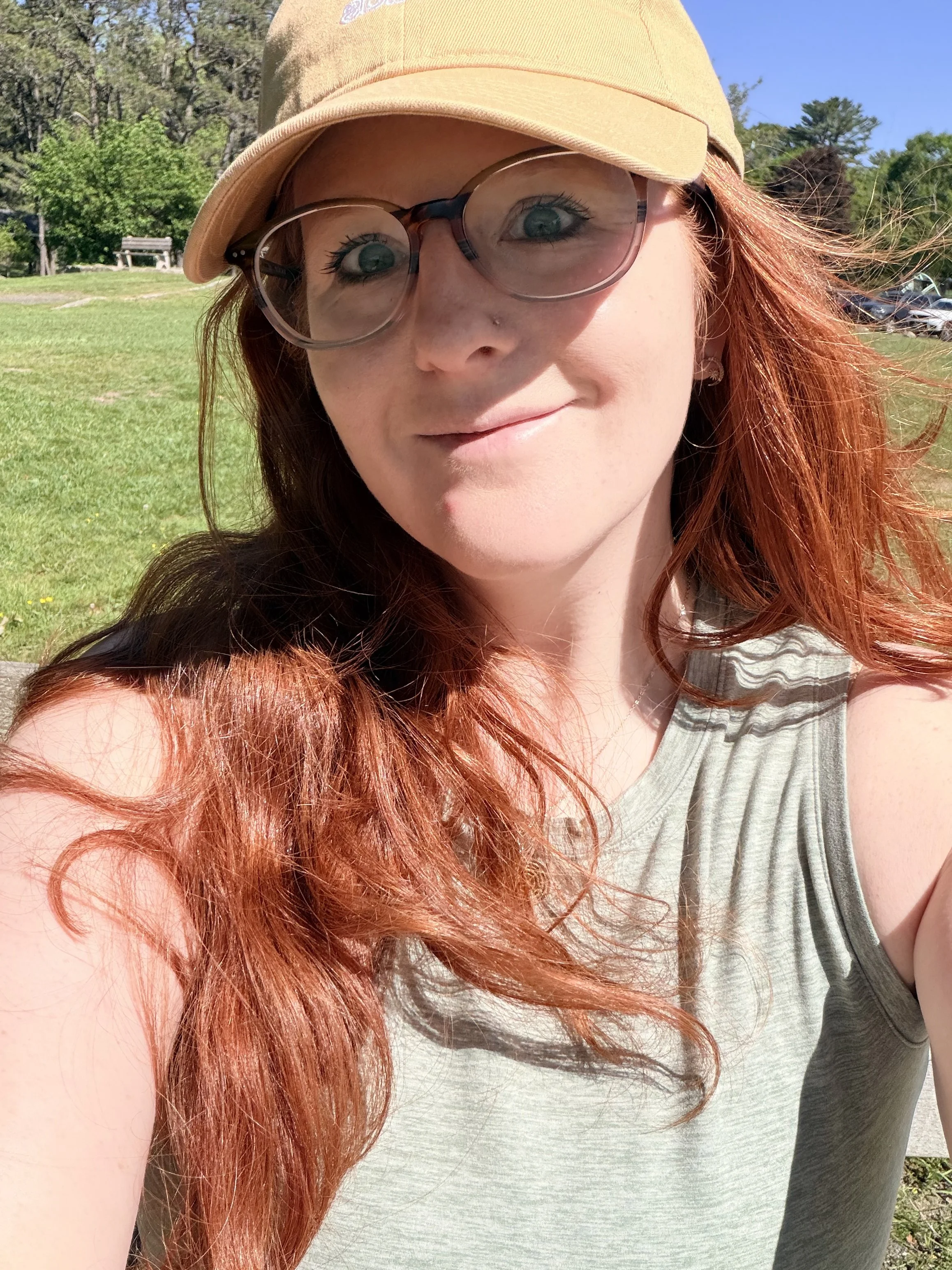 A woman with long red hair wearing glasses, a tan cap, and a gray tank top taking a selfie outdoors on a sunny day with trees and a parking lot in the background.
