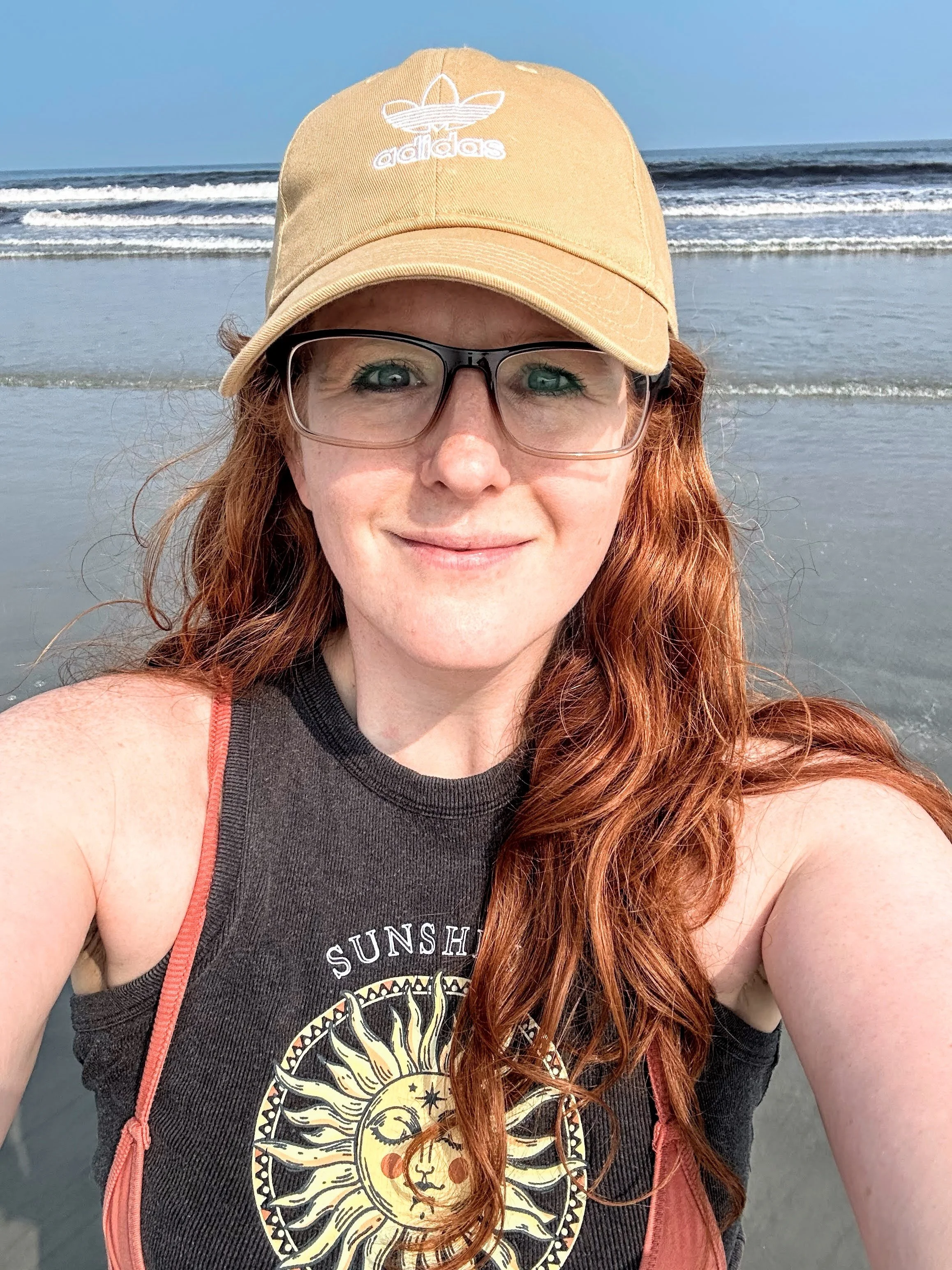 A woman with long red hair, wearing glasses, a beige Adidas cap, a black sleeveless shirt with a sun and moon graphic, and a reddish-orange strap, taking a selfie at the beach with ocean waves and blue sky in the background.