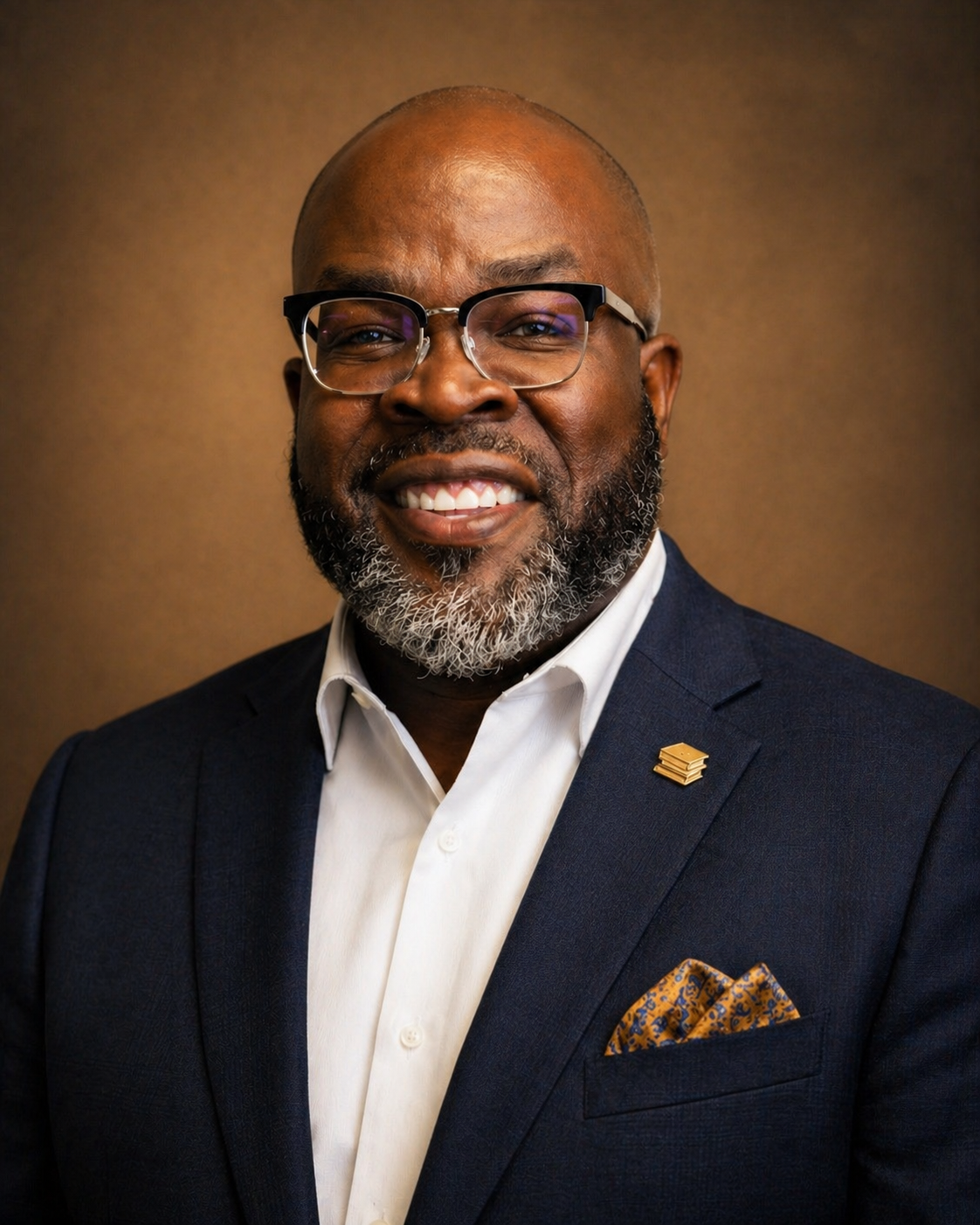 A man with glasses, a beard, and a bald head wearing a dark suit jacket, white shirt, and a patterned pocket square. He is smiling and standing against a brown background.