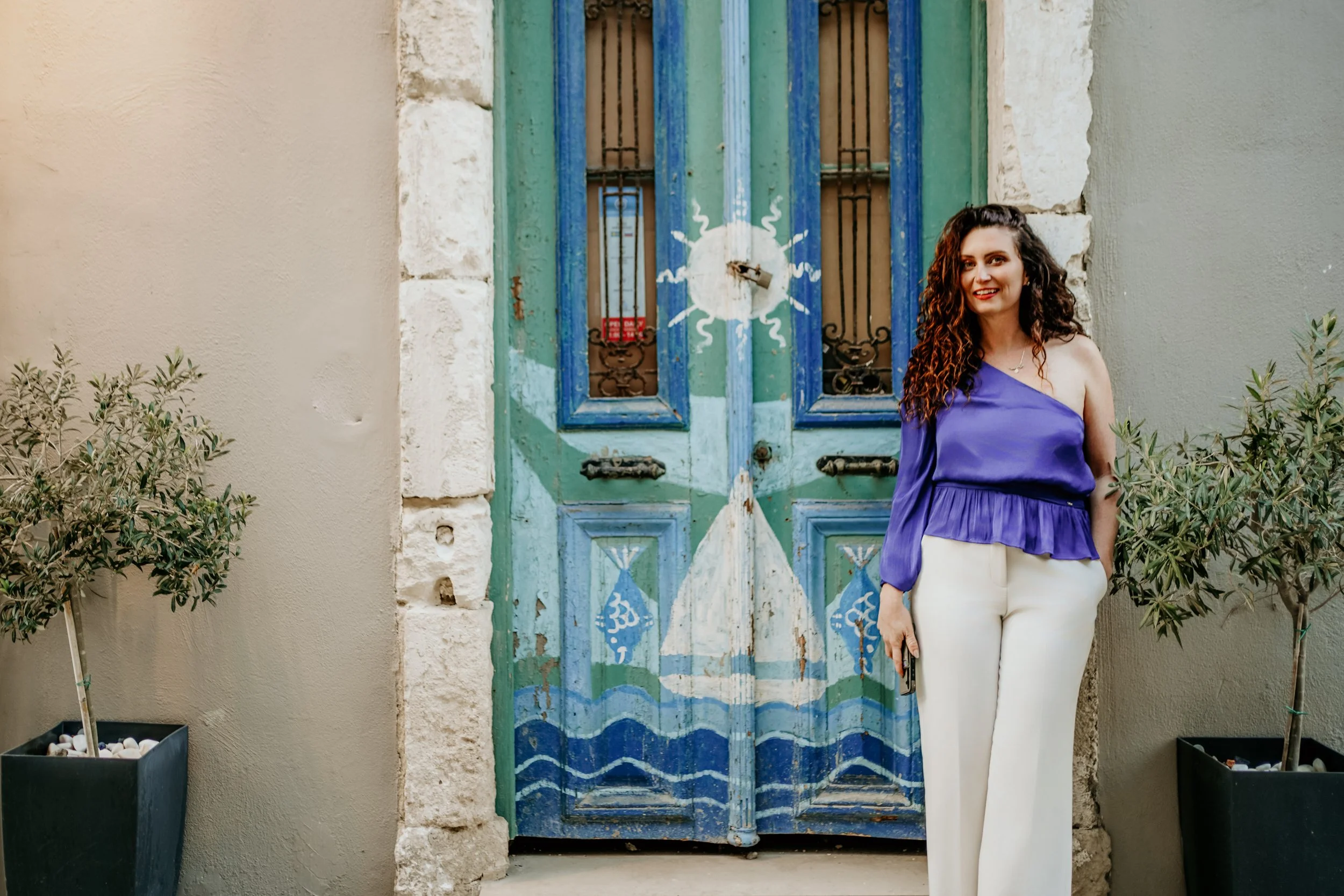 Cristina Nicole posing by a vibrant Greek doorway framed with lush potted plants and Mediterranean charm.