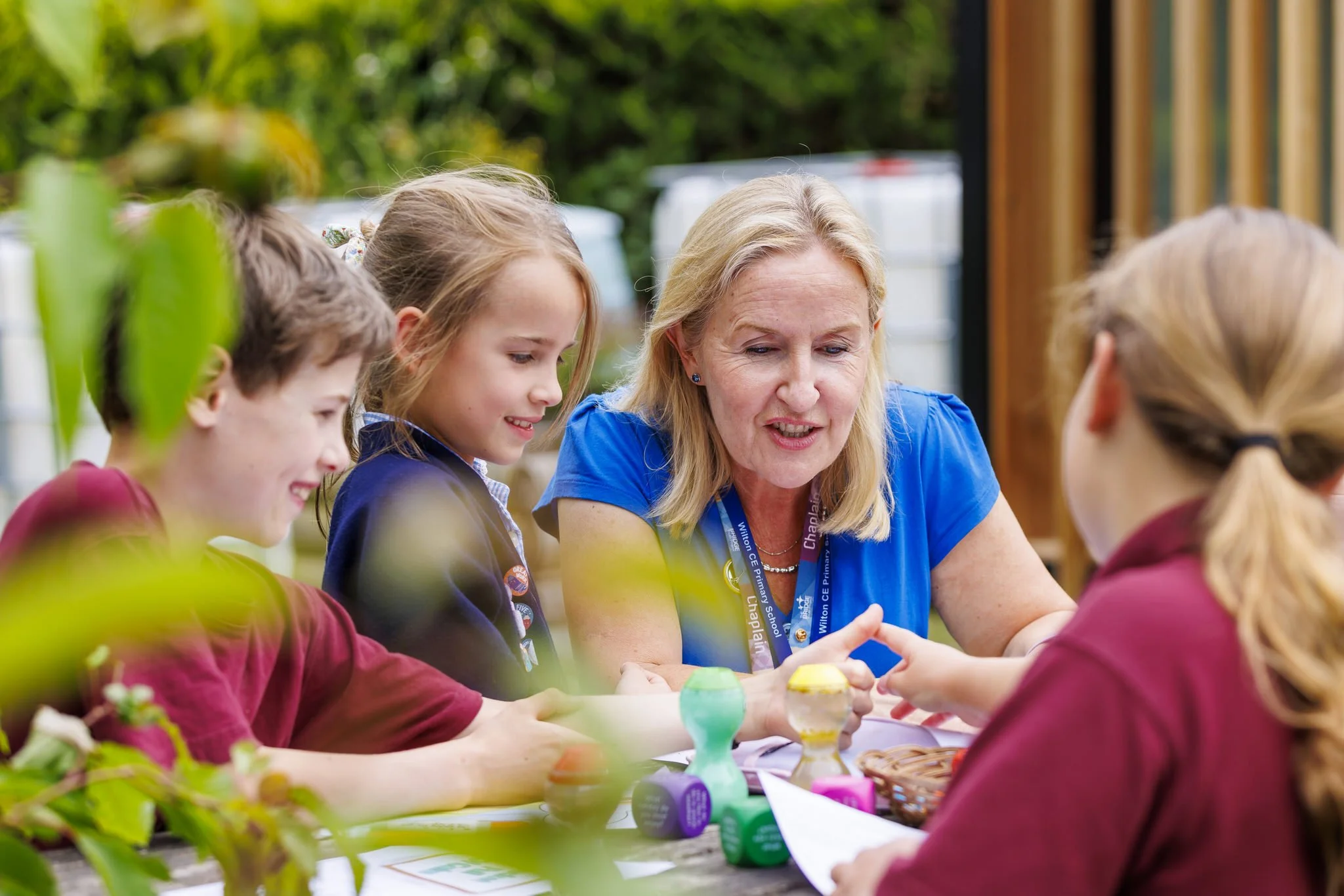 A teacher or adult woman engaging with children at an outdoor craft or activity table, surrounded by greenery, with colorful craft supplies on the table.