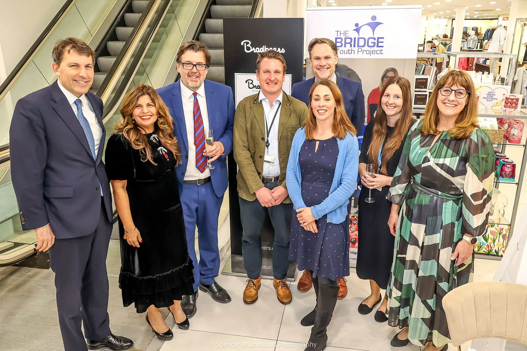 A group of eight well-dressed people, four men and four women, standing together smiling in a shopping mall near escalators and promotional banners for The Bridge Youth Project and Bradbeers, celebrating a social or community event with drinks in hand.