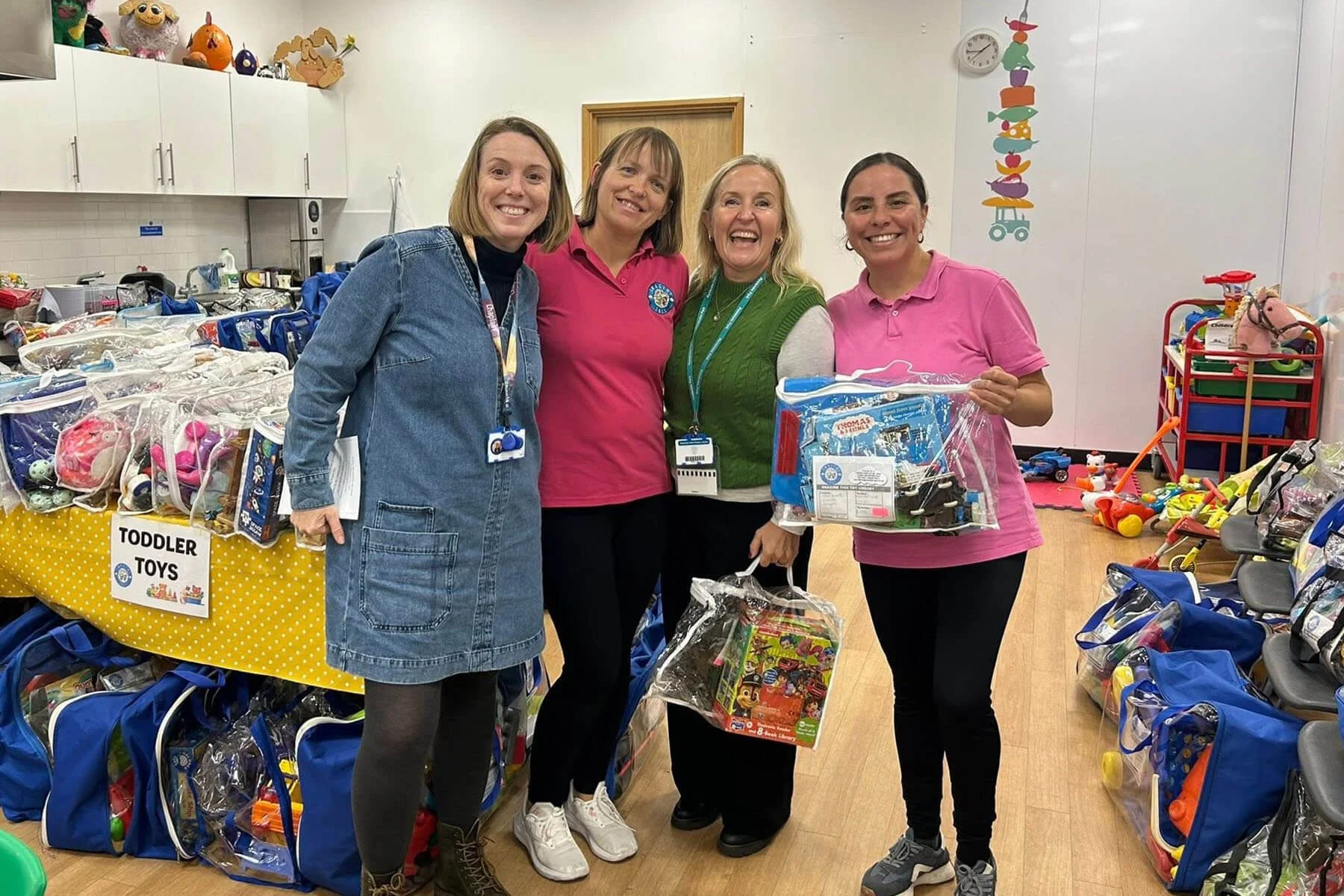Four women standing in a room filled with donated toddler toys and children's items, smiling at the camera.