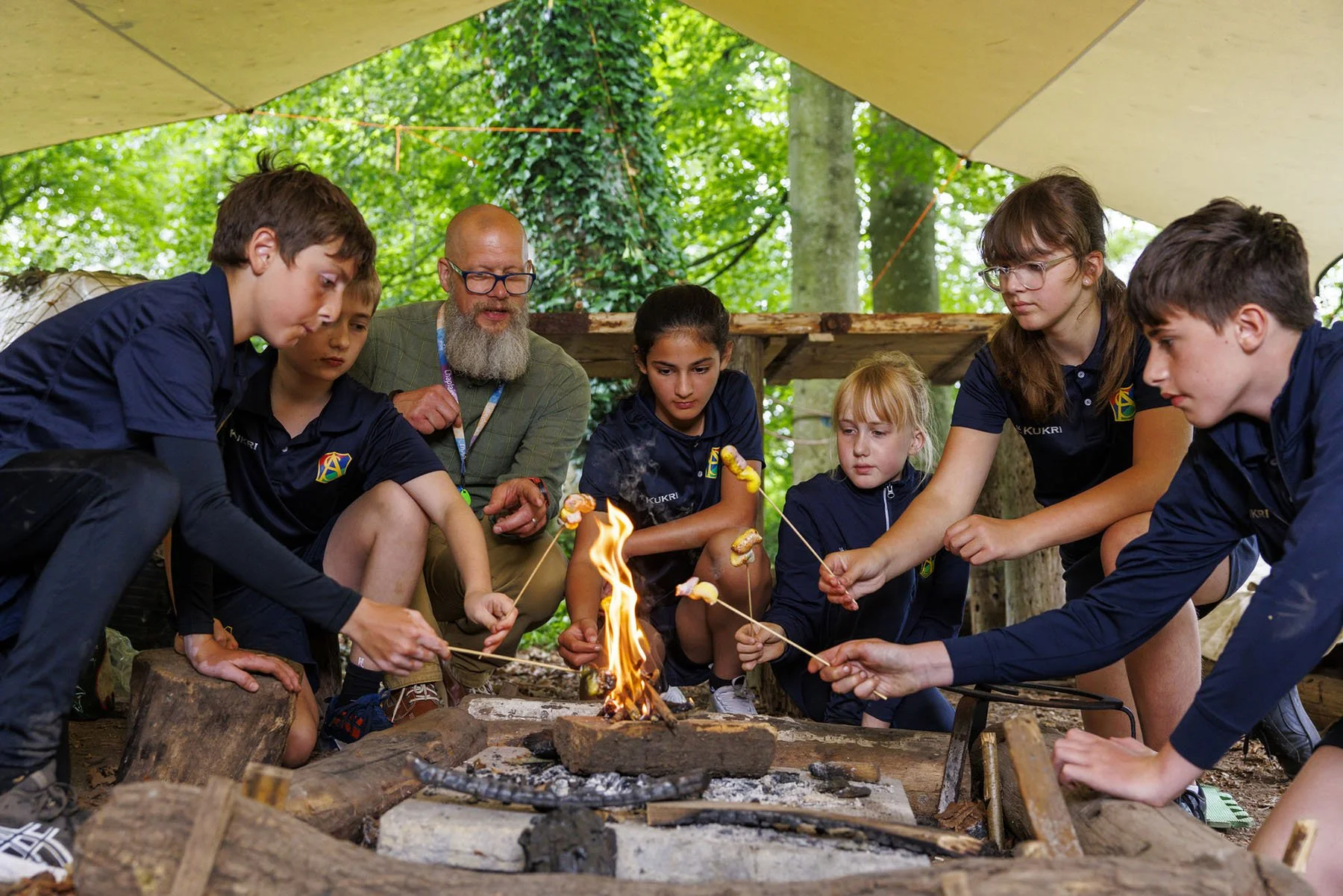 A group of children and a man gathered around a campfire in a wooded outdoor setting, roasting marshmallows on sticks.