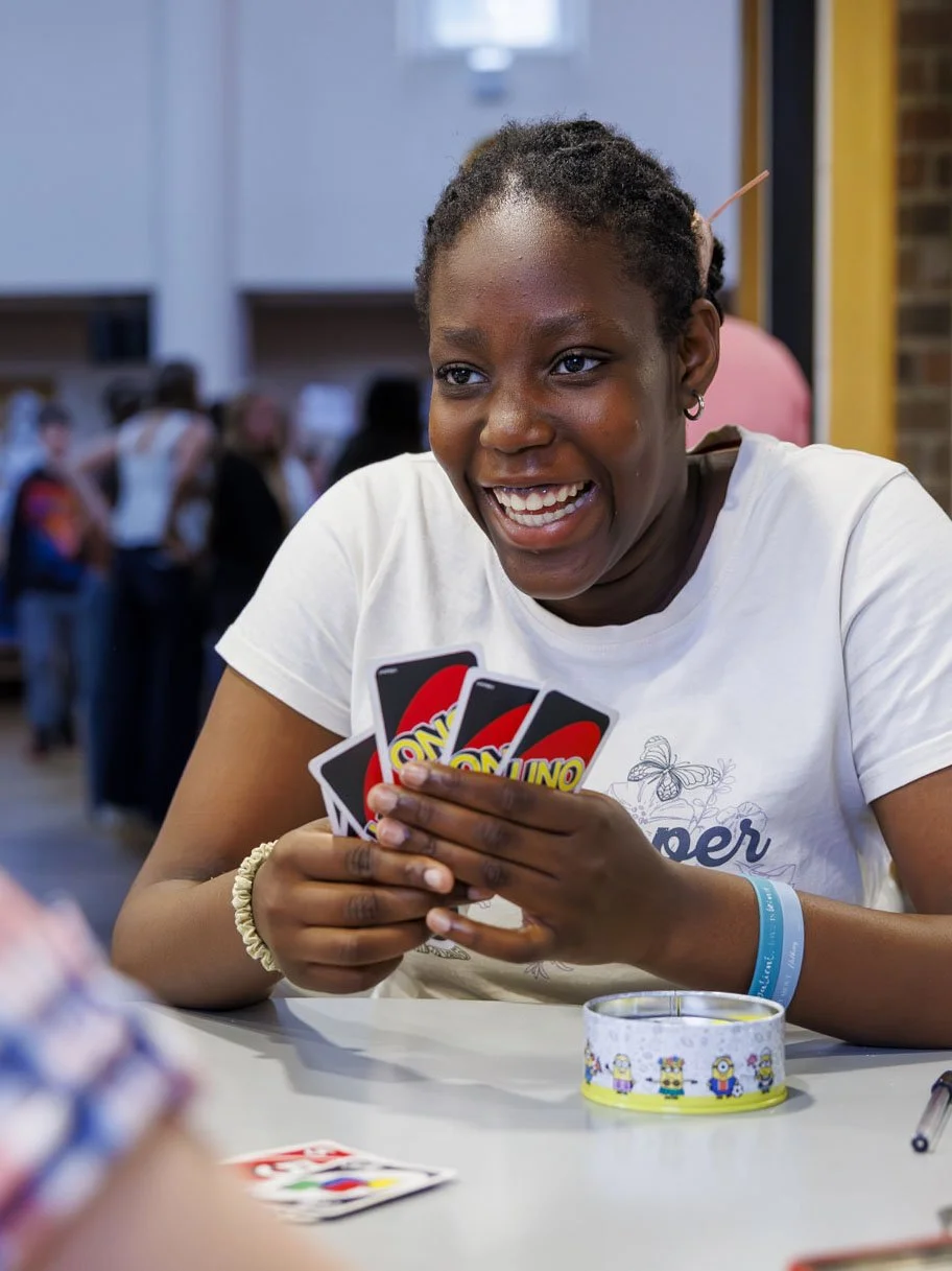 A young woman smiling widely while holding Uno cards at a table, with a roll of Minions-themed tape and a marker in front of her, in a lively indoor setting.
