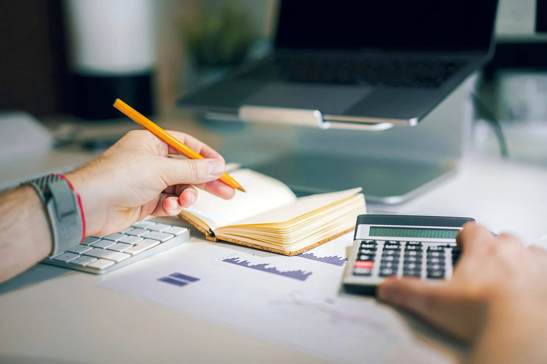 A person with a wristwatch and a red bracelet taking notes with a yellow pencil on a notepad at a cluttered desk. The desk also has a computer keyboard, a calculator, printed charts or graphs, and a laptop with a document holder in the background.