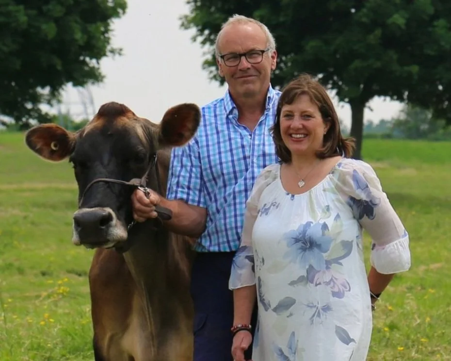 A man and woman standing outdoors in a grassy field with trees in the background, along with a brown cow with a halter, the man holding the cow's halter.