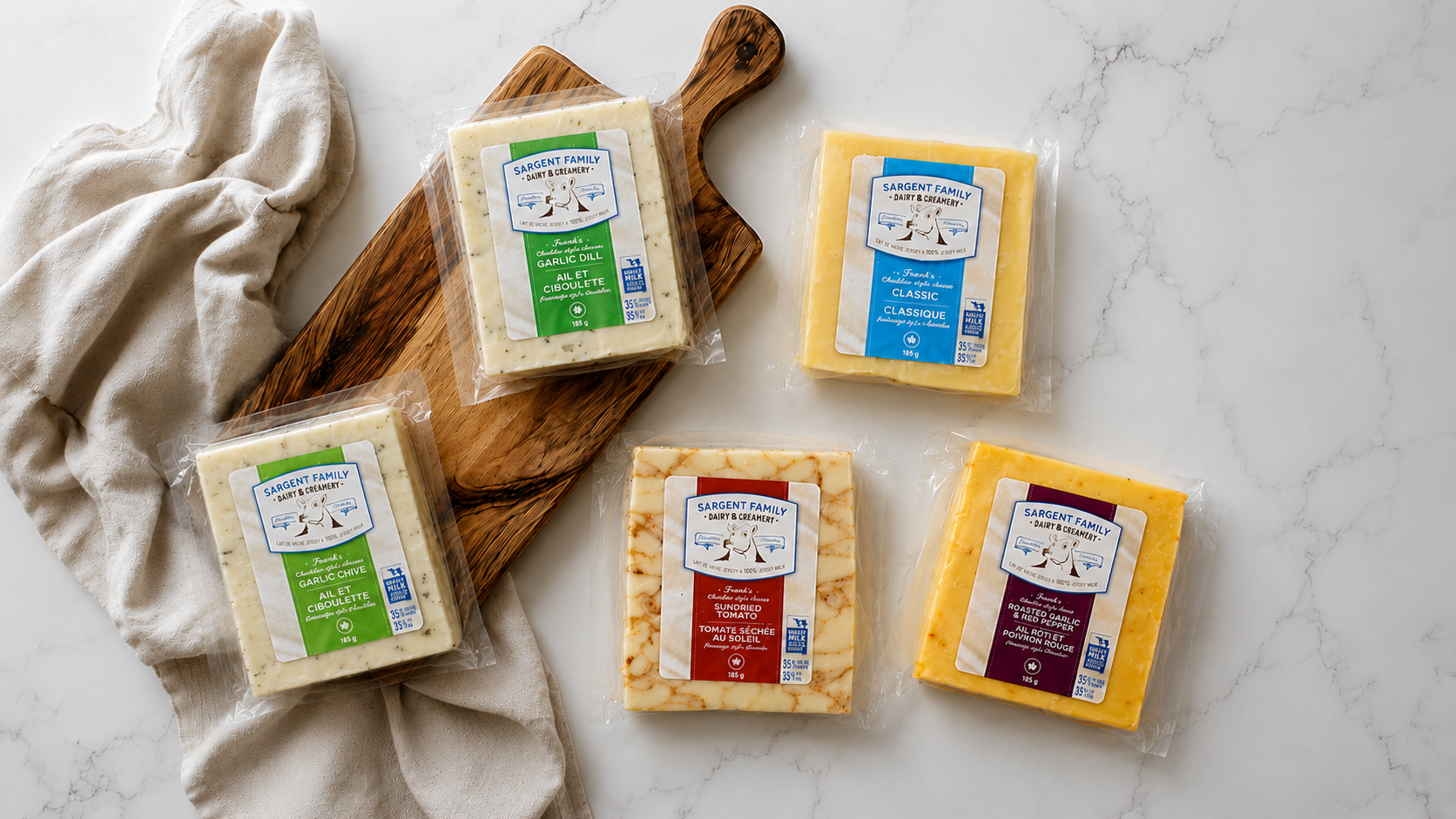 Assorted blocks of cheese in packaging on a white marble surface, with a wooden cutting board and a cream-colored cloth.