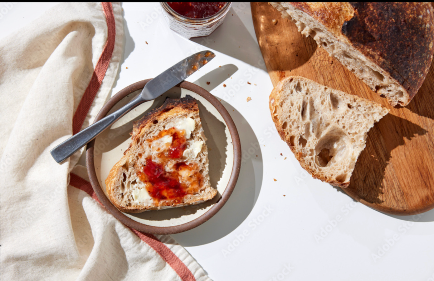 Slice of crusty bread with butter and jam on a small plate, bread on a cutting board, and a jar of jam.