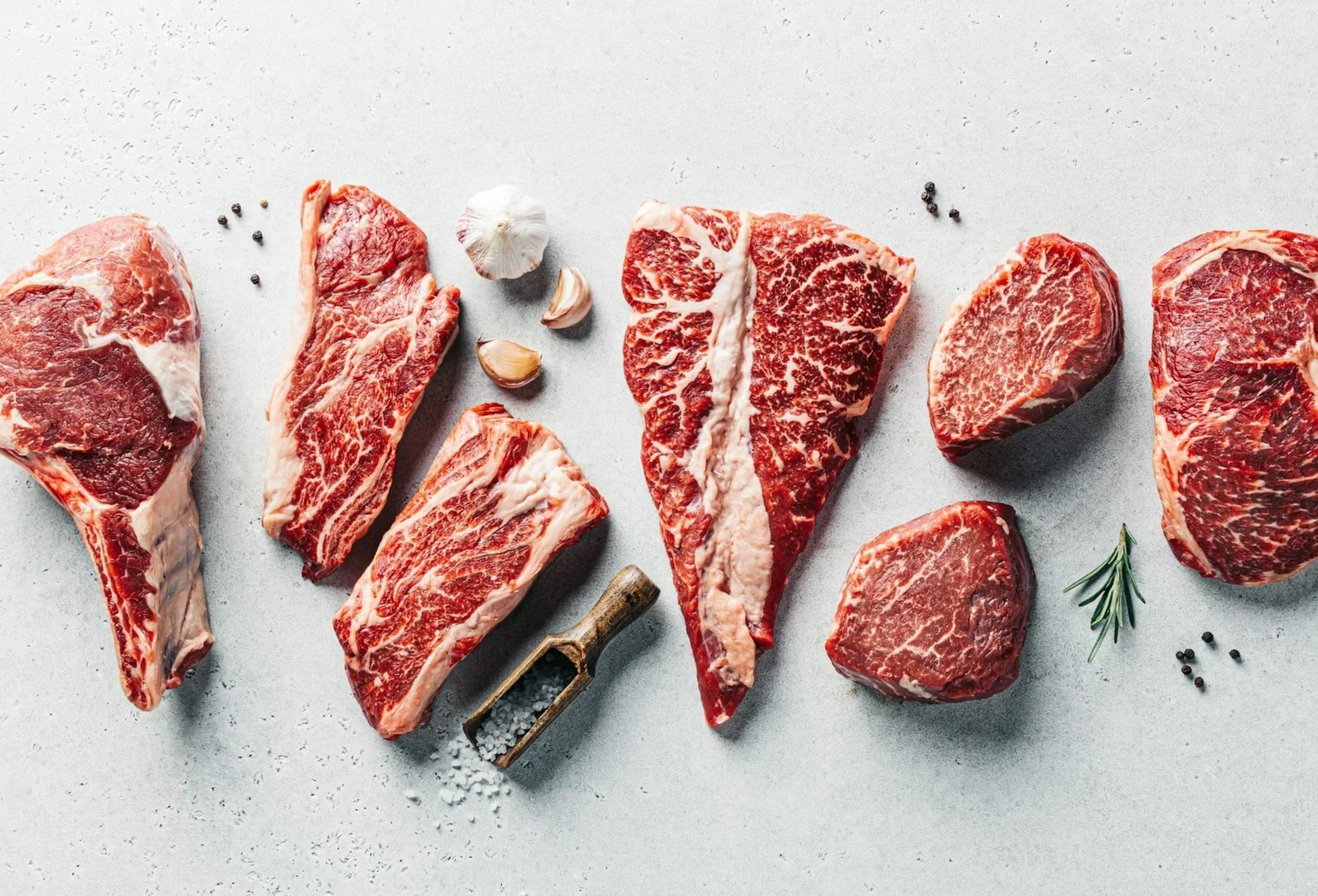 Several raw beef steaks with marbling, garlic cloves, black peppercorns, sea salt, and a sprig of rosemary on a light gray surface.