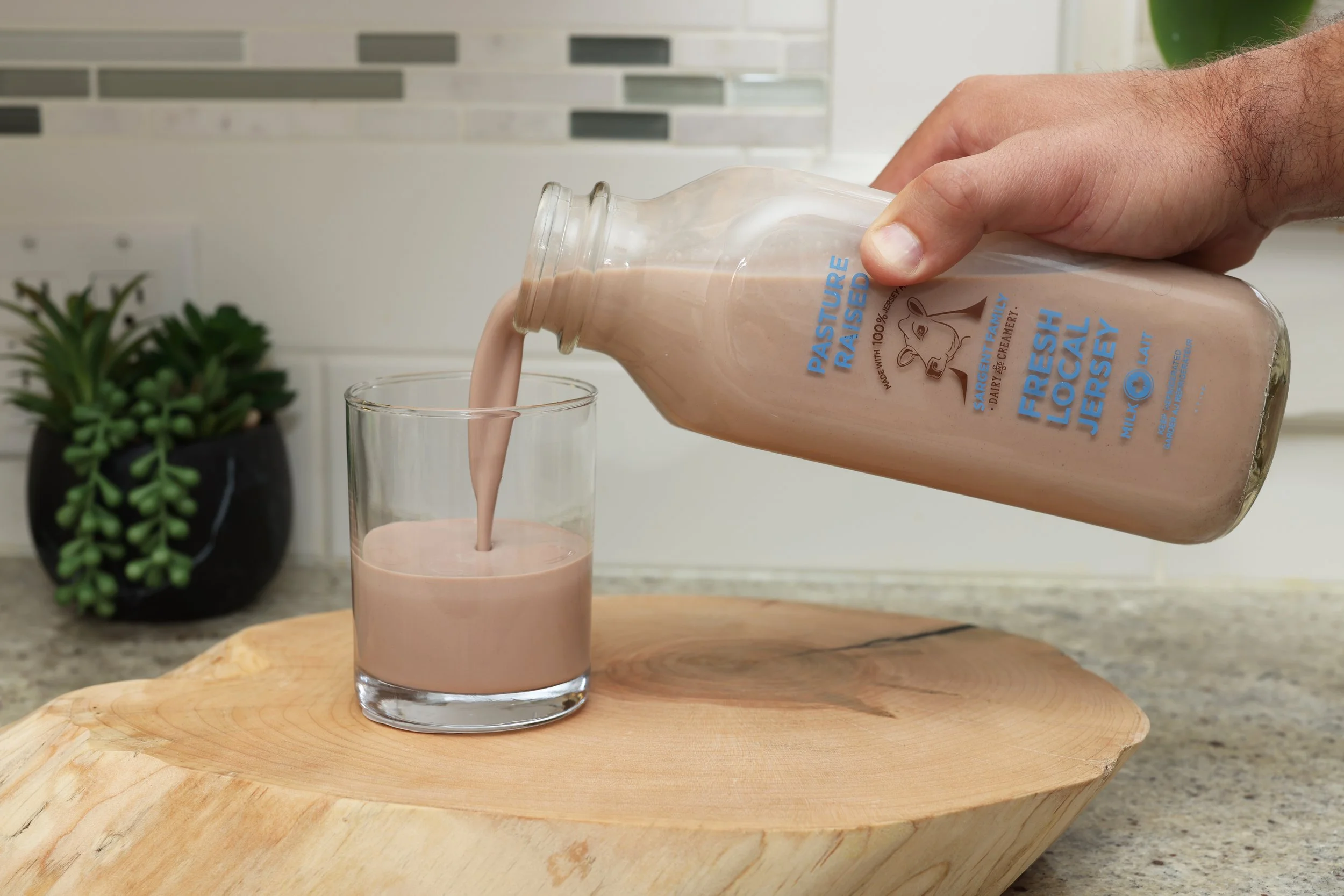 A person pours chocolate milk from a bottle into a glass on a kitchen countertop, with a small plant in the background.