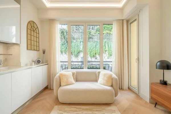Bright living room with a cream sofa, large window with curtains, and minimal decor including a black lamp on a wooden side table.