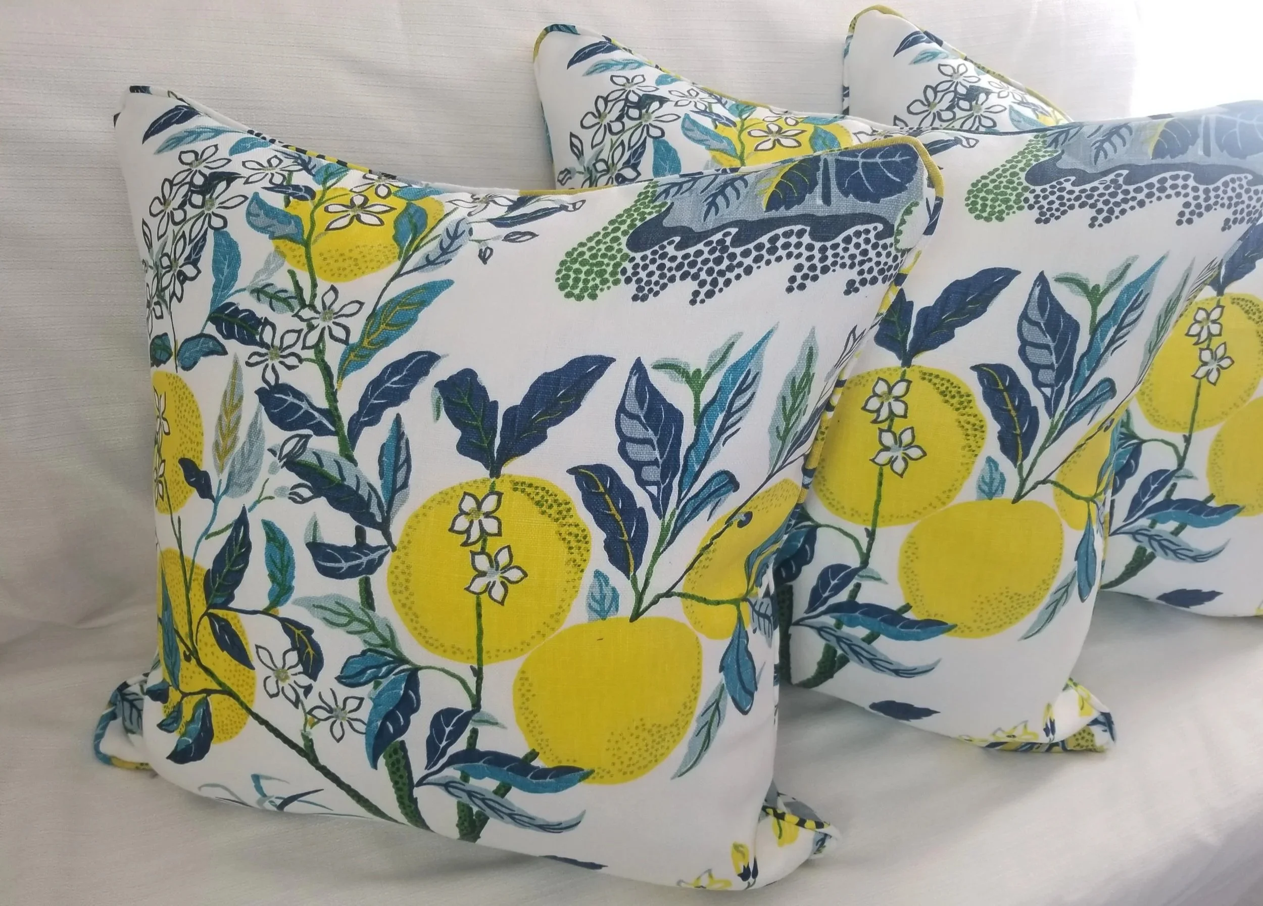 Four decorative pillows with a floral and lemon print in blue, green, yellow, and black on a white background, arranged on a bed or sofa against a light-colored wall.