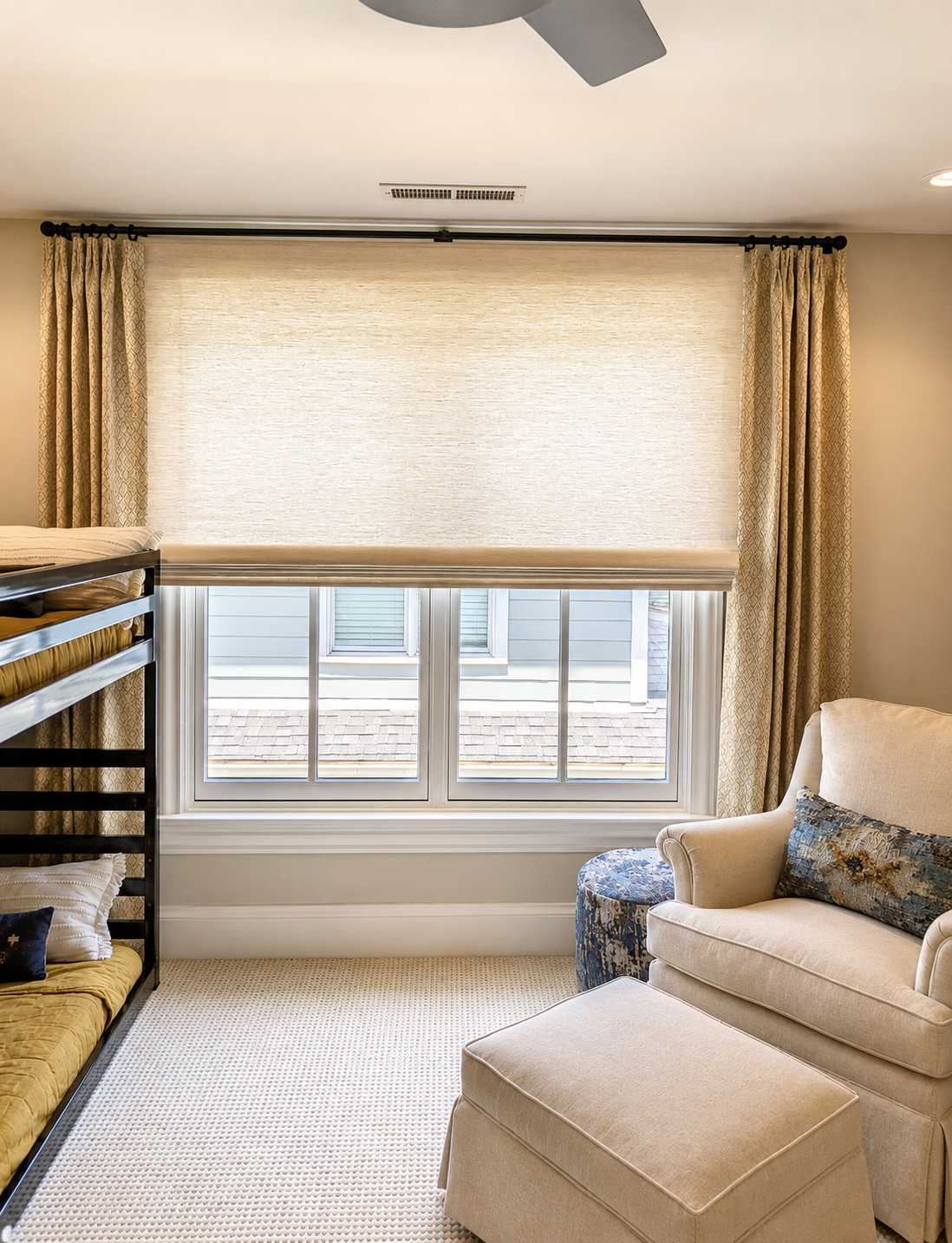 A room with a large window covered by a beige roller shade, flanked by patterned curtains. Part of a black bunk bed and a beige armchair with ottoman are visible.