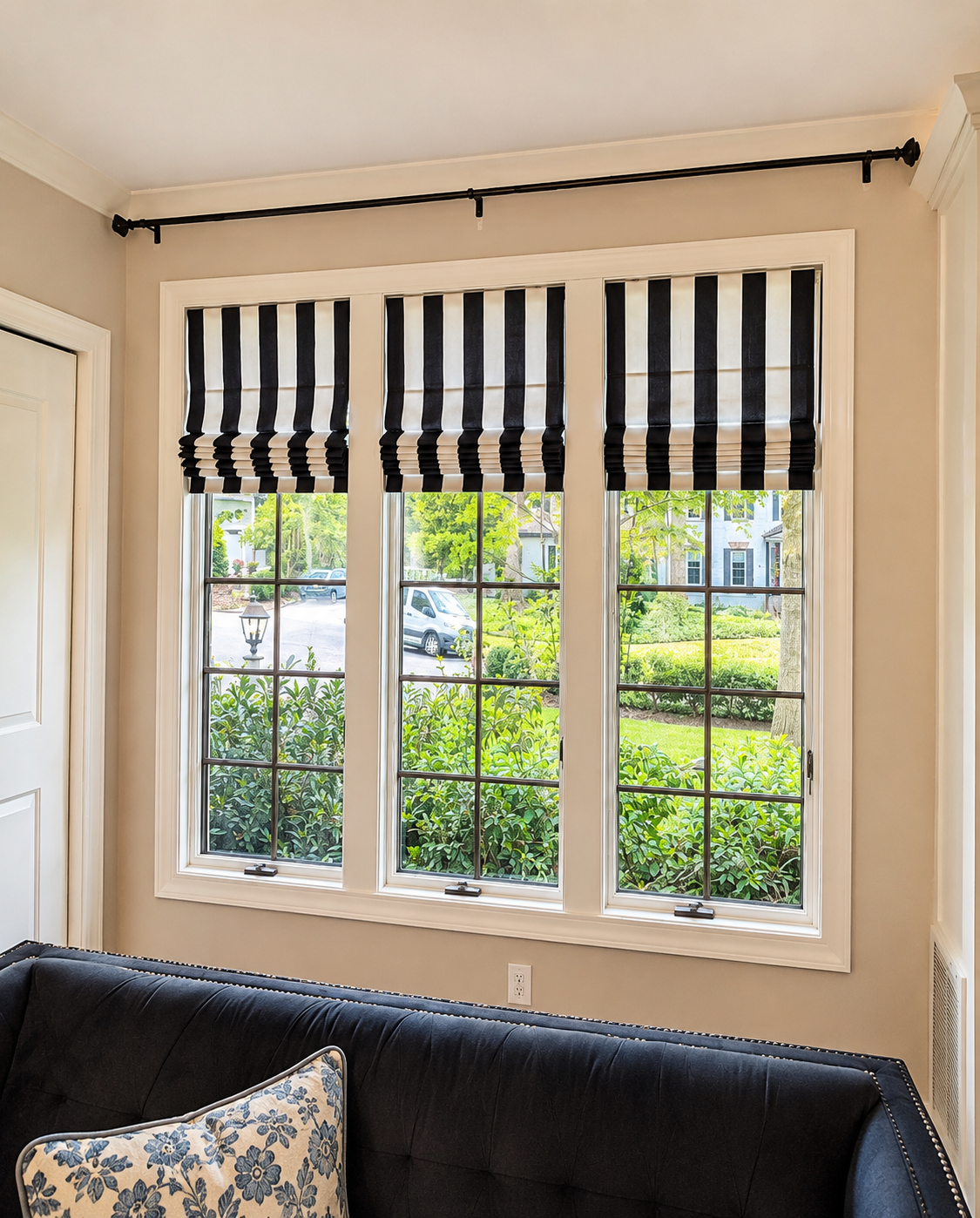 Three large windows with black and white striped Roman shades, overlooking a front yard with greenery and parked cars.