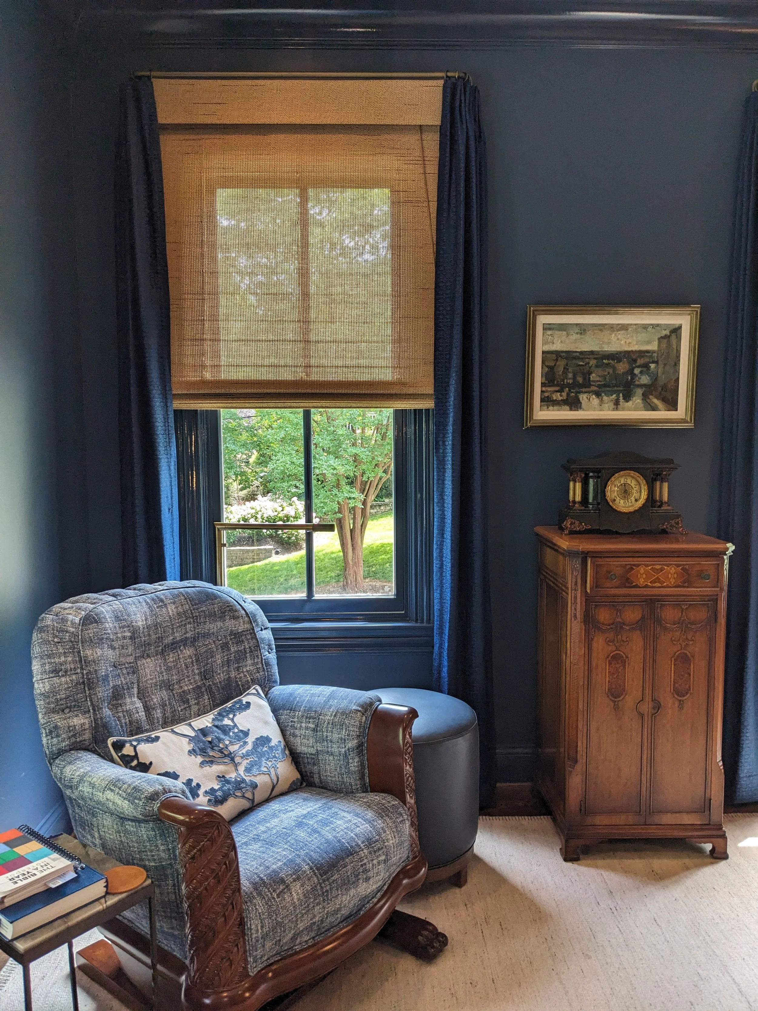 A living room corner featuring a blue wall, a window with beige shade and blue curtains, a plaid armchair with wooden arms and a decorative pillow, a small side table with books, a vintage wooden clock cabinet, a framed landscape painting, and a beig