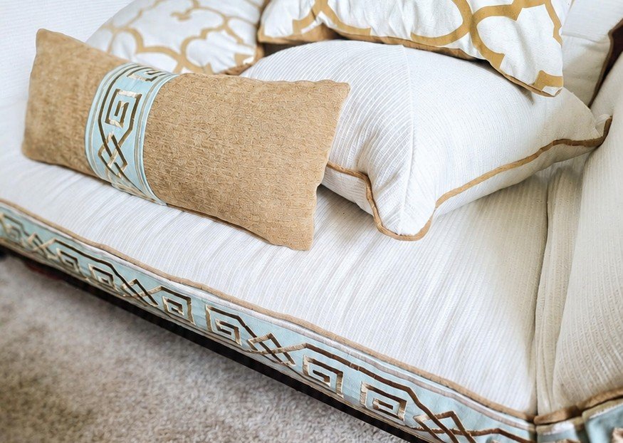 A bed with white sheets, a decorative blue and beige trim on the mattress, and several pillows including a beige bolster pillow with a blue ribbon and two white pillows with beige and gold patterns.
