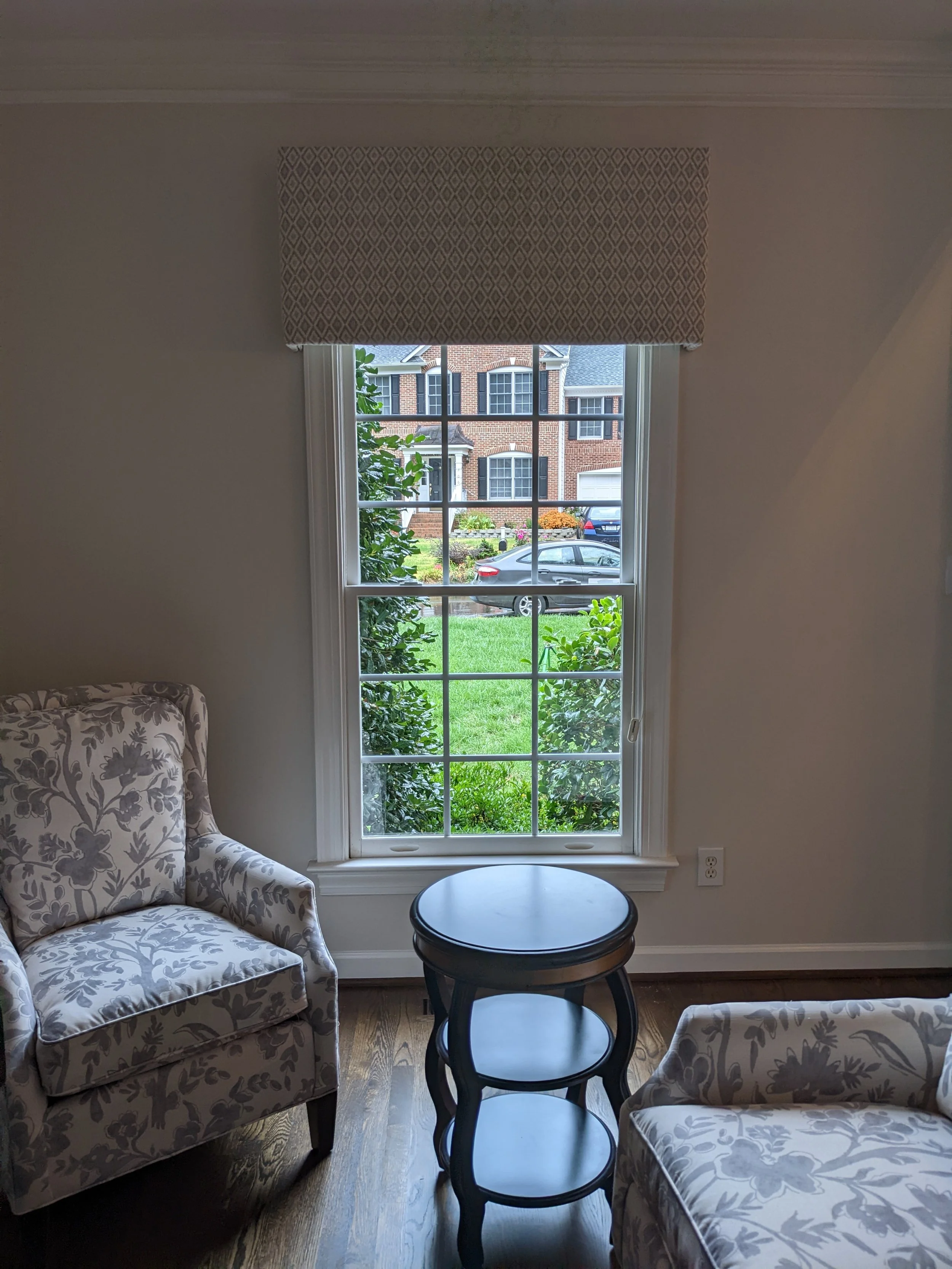 Interior view of a room with a large window showing a neighborhood street, two armchairs with floral pattern, and a small round table.
