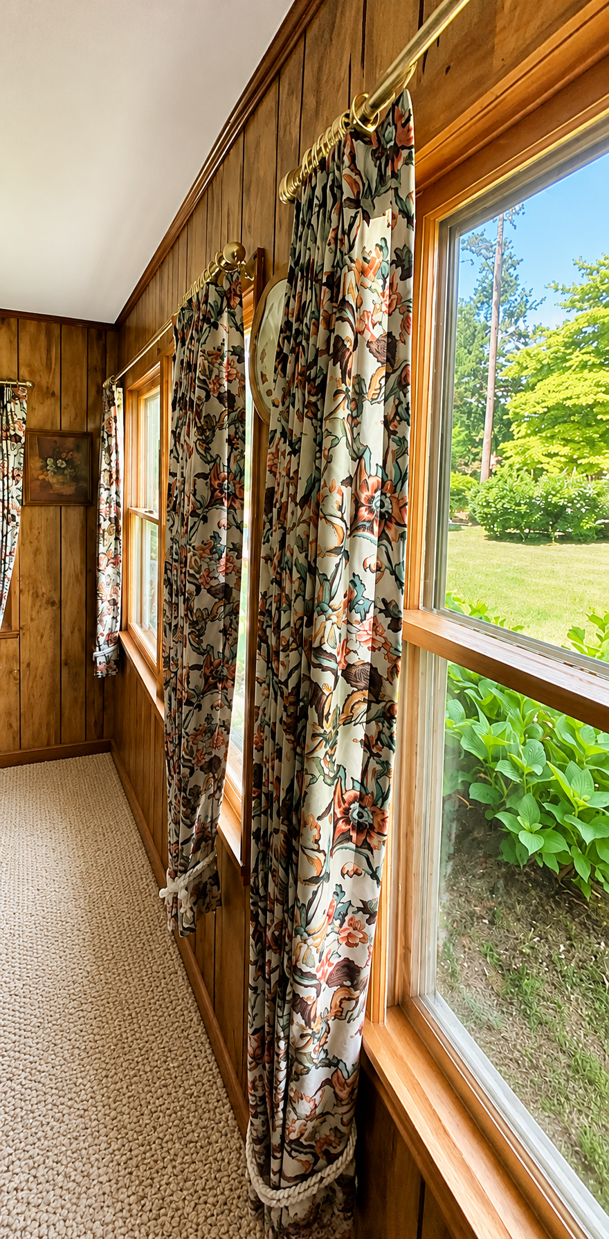 View of a wood-paneled room with floral curtains hanging from a brass curtain rod and windows showing a garden outside.