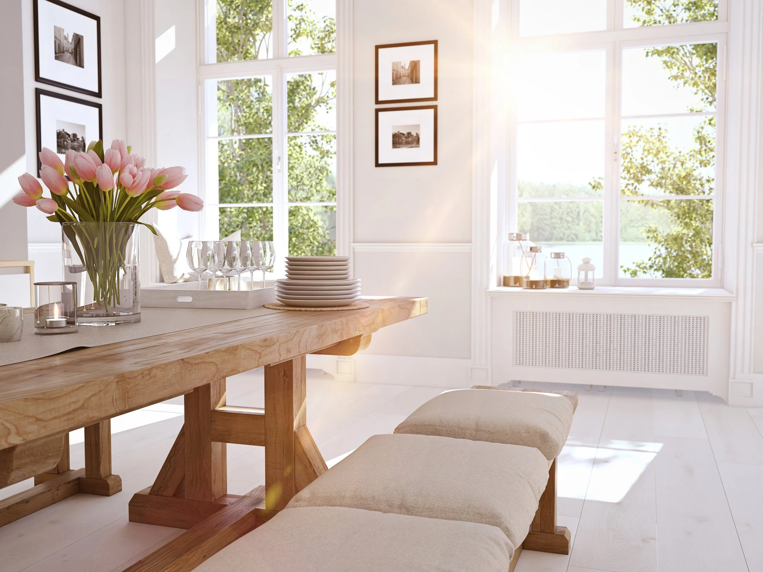 A bright dining area with a wooden table set with a vase of pink tulips, glasses, and stacked plates. Large windows let in natural light and show green trees outside. Framed black and white photographs are on the white walls.