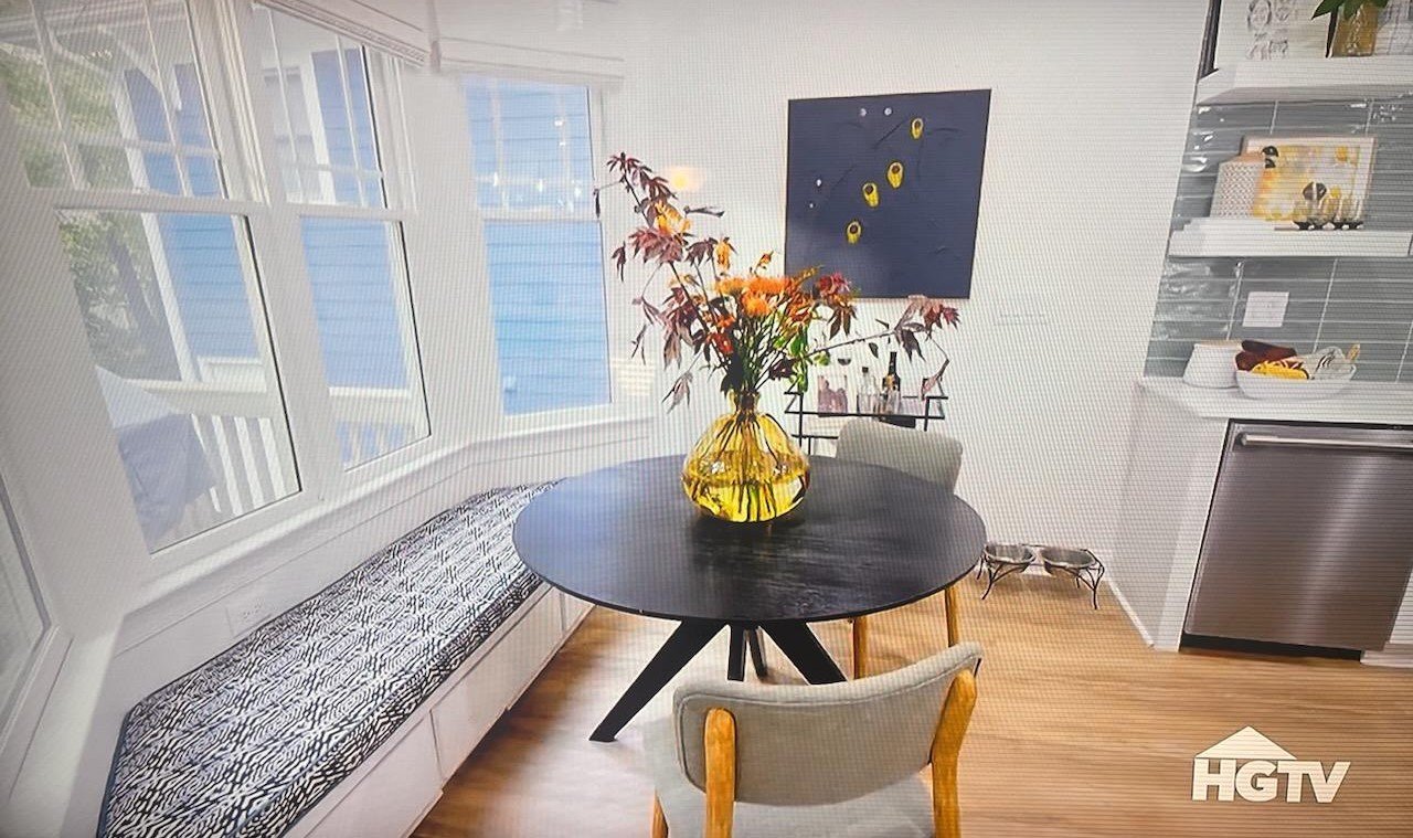 Round black dining table with a yellow glass vase holding branches and leaves, surrounded by chairs, in a bright room with large windows, a bench with animal print cushion, and a kitchen area in the background.