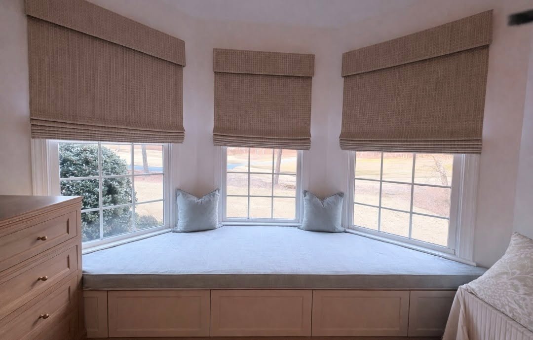 A cozy window nook with a cushioned seat, three large windows with beige blinds, and two pillows on the window seat.