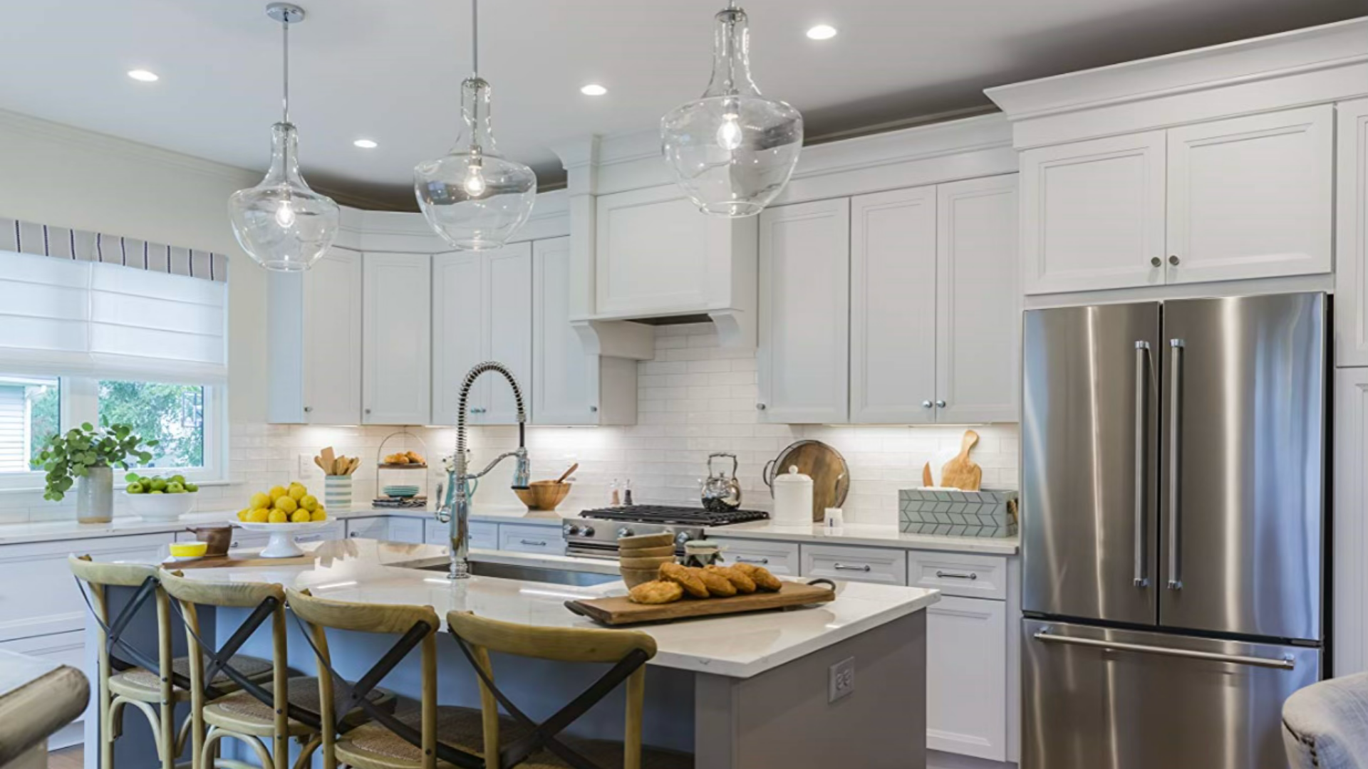 Modern white kitchen with island, pendant lights, stainless steel refrigerator, and window with blinds. Countertop has lemons, green apples, and baked goods.