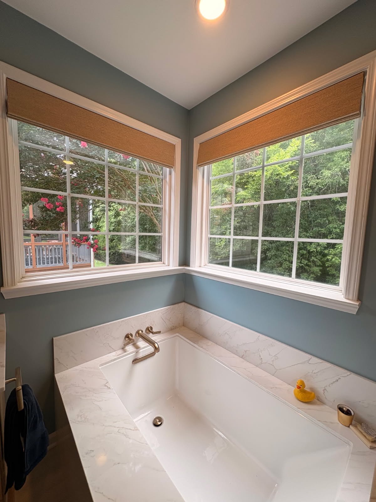 Bathtub with marble surround and two large windows showing greenery outside; a small rubber duck on the edge of the bathtub; a towel hanging on a hook on the left side.