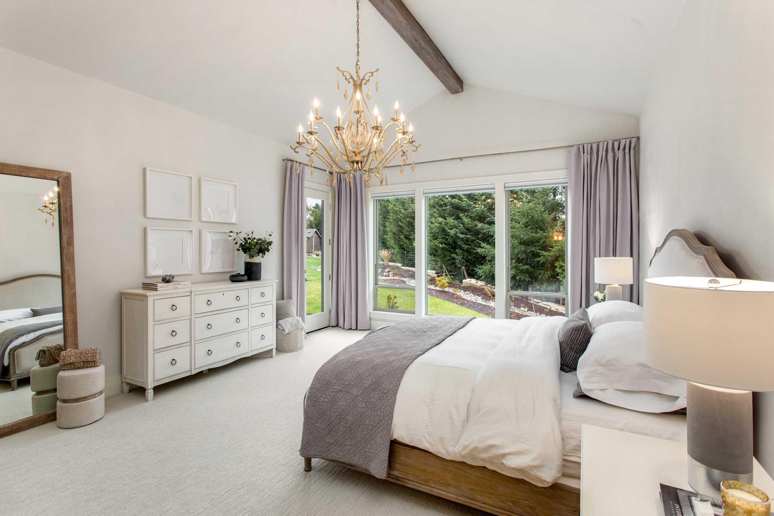 A bedroom with a large window showing trees outside, a chandelier hanging from the ceiling, a bed with white bedding and dark gray blanket, white nightstand with a lamp, a white dresser with decorative items, a full-length mirror, and light curtains.