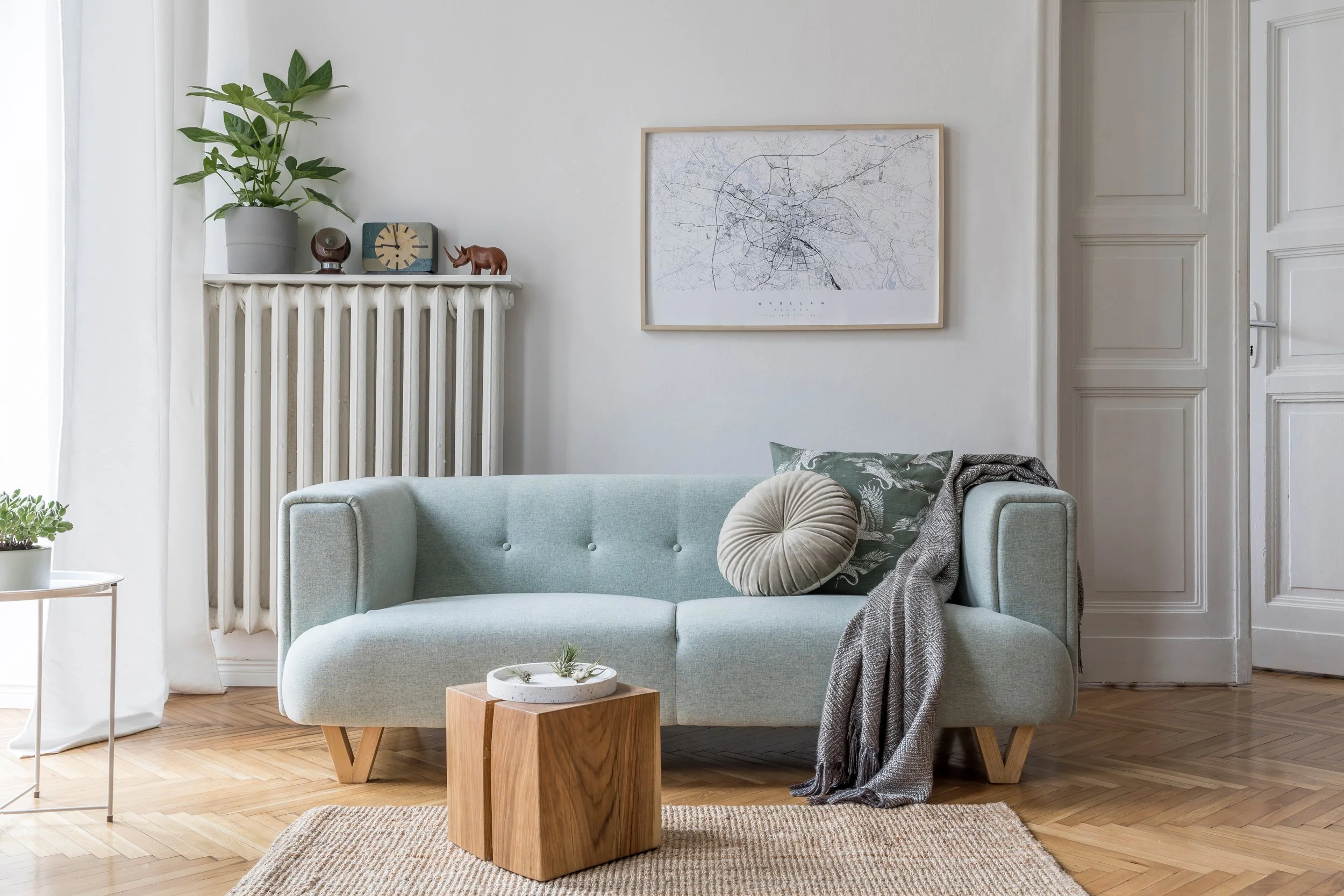A cozy living room with a light blue sofa, round pillow, a throw blanket, a wooden coffee table with small plants, and a side table with a potted plant. The wall behind features a large framed city map, a white radiator decorated with a large plant and small decorative items, and a closed paneled door.