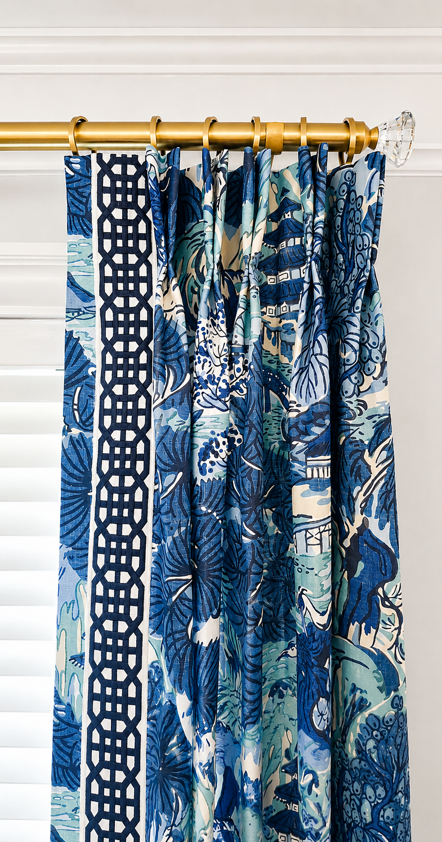 Patterned blue and white curtains hanging on a gold curtain rod with a clear glass finial, partially covering a window with white Venetian blinds.
