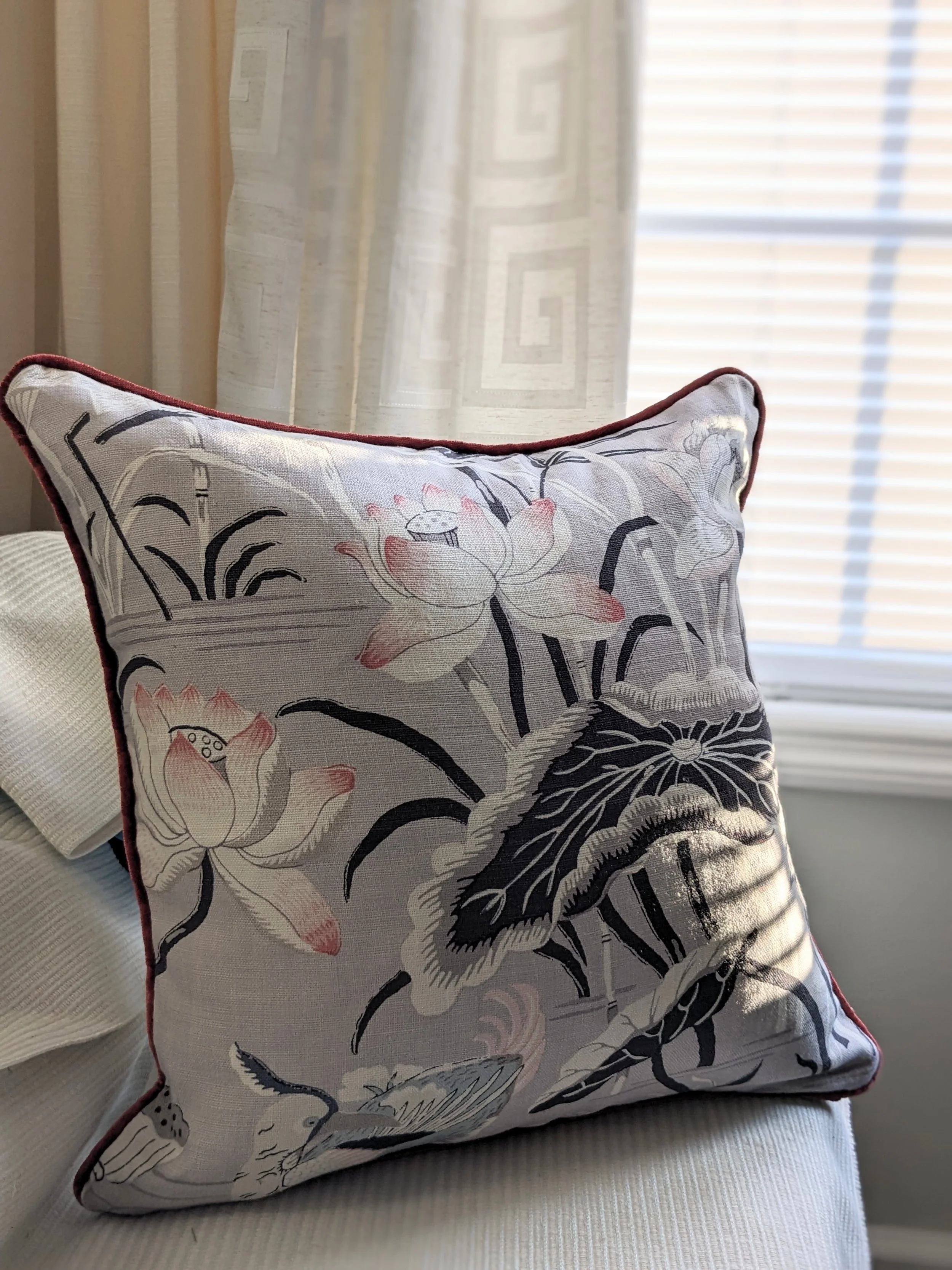Close-up of decorative pillow with floral and animal design on a fabric sofa, lightly illuminated by sunlight through window blinds, with sheer curtains in the background.