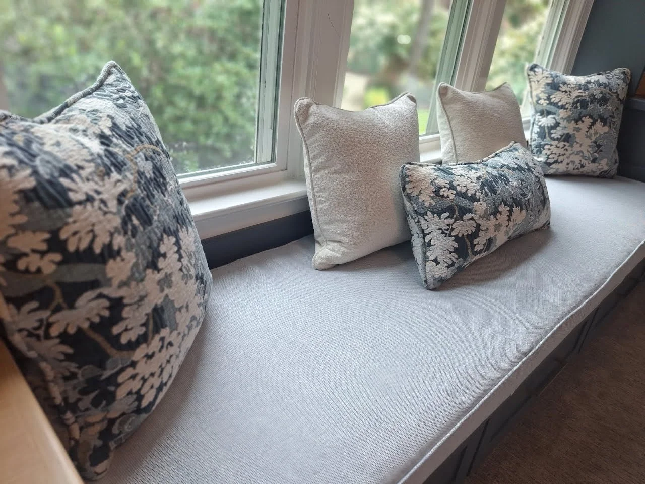 Window seat with four decorative pillows, two floral patterned and two plain, facing a large window with greenery outside.