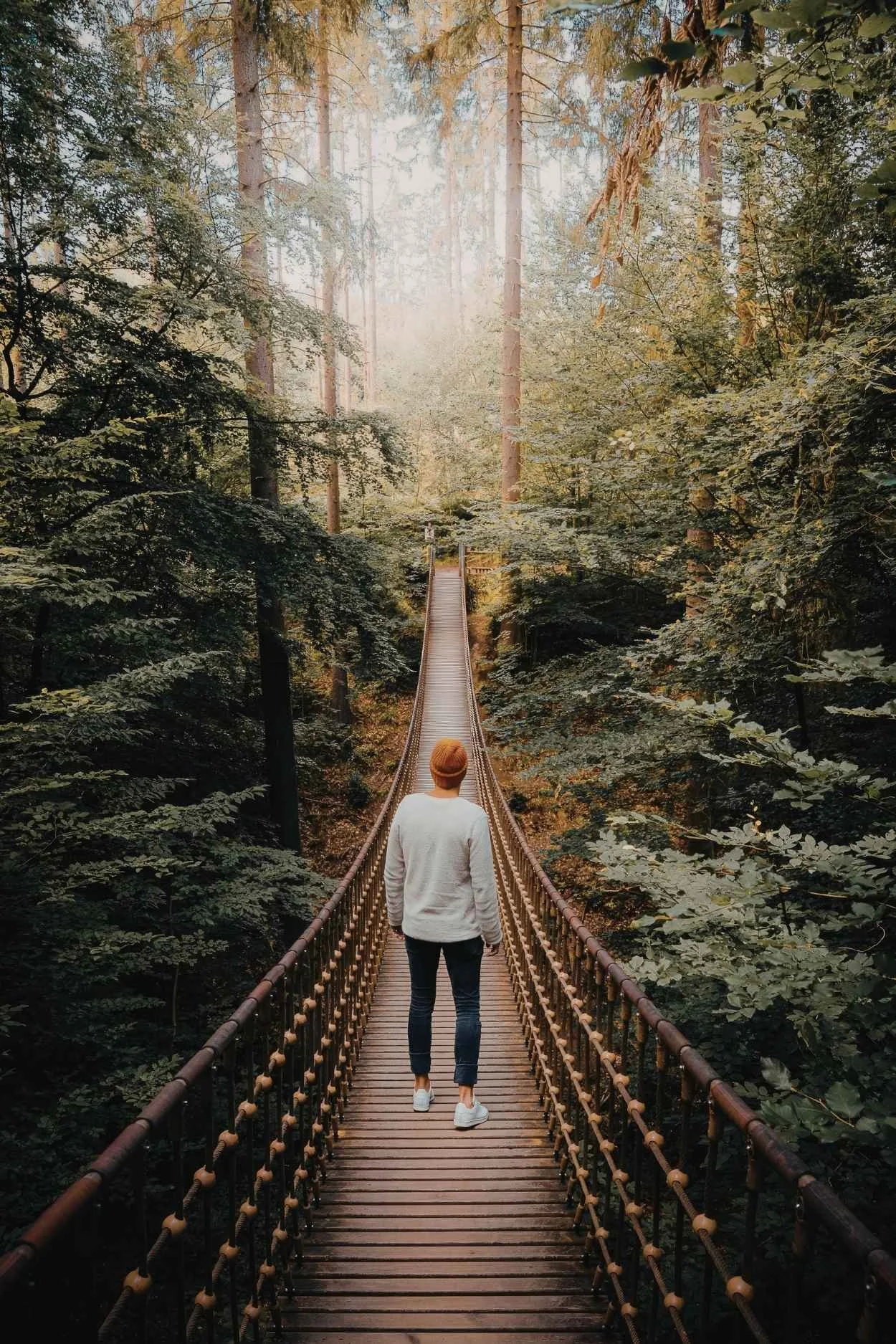 A person in a white sweater, dark pants, and white shoes walking across a wooden suspension bridge in a dense forest with tall trees and sunlight filtering through the leaves.
