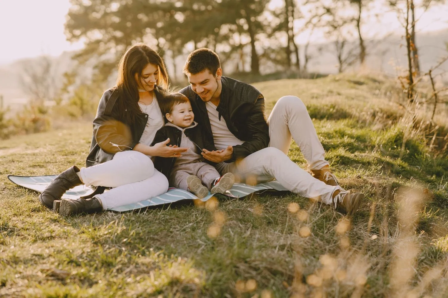 A family of three sitting on a blanket outdoors during sunset, smiling and looking at a smartphone together.