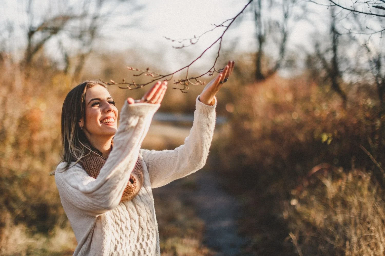 A woman in a beige sweater and a brown scarf smiling and reaching up to touch a bare tree branch during autumn.