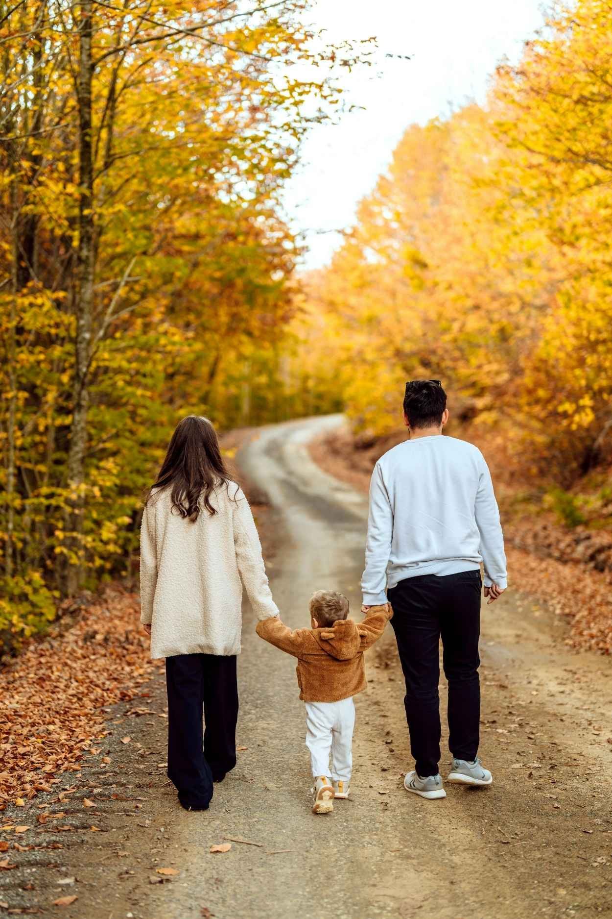 A family of four walking along a dirt path in a forest with colorful autumn trees on both sides.