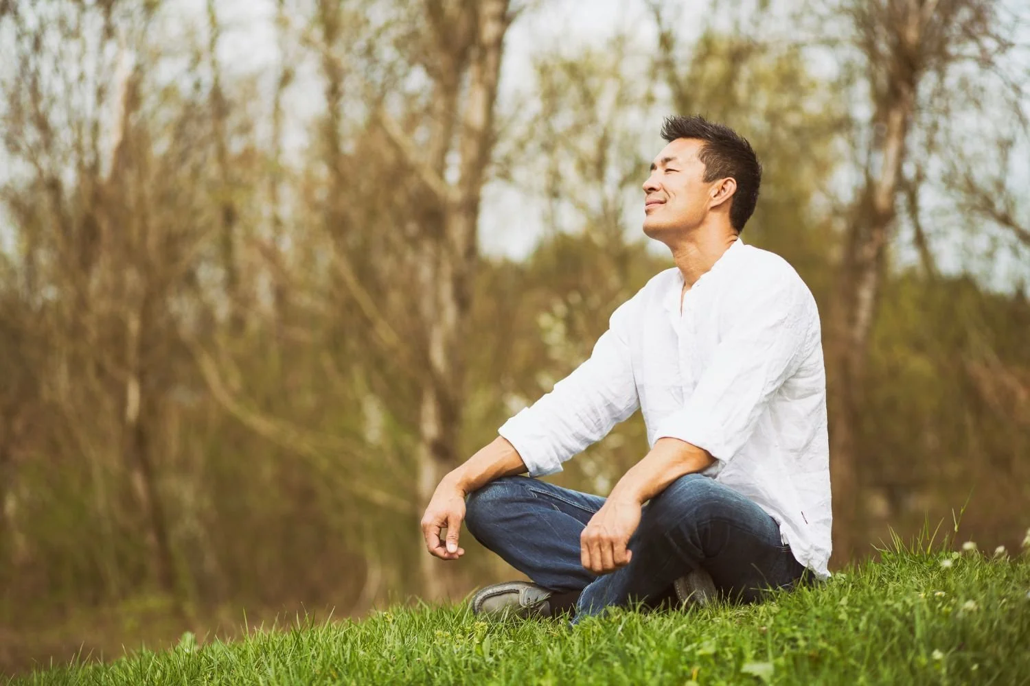 A man sitting cross-legged on grass in a peaceful outdoor setting, smiling with eyes closed, enjoying nature with trees in the background.