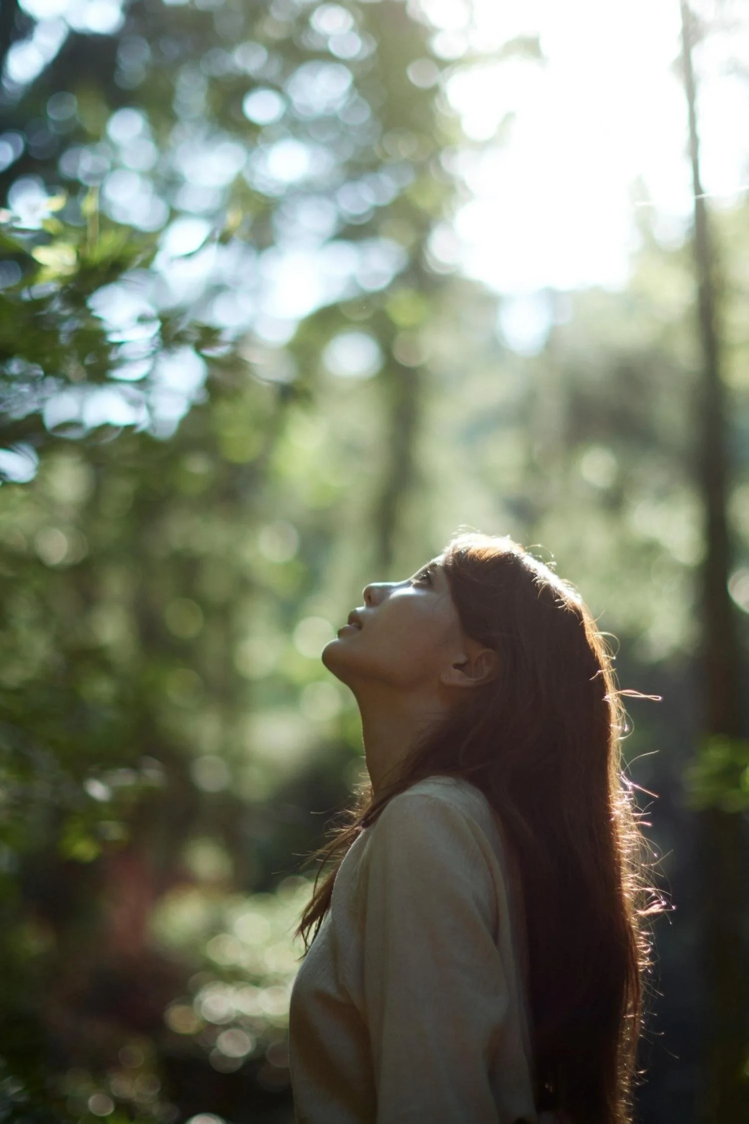 A young woman with long red hair standing outdoors in a forest, looking up towards the bright sunlight filtering through the trees.