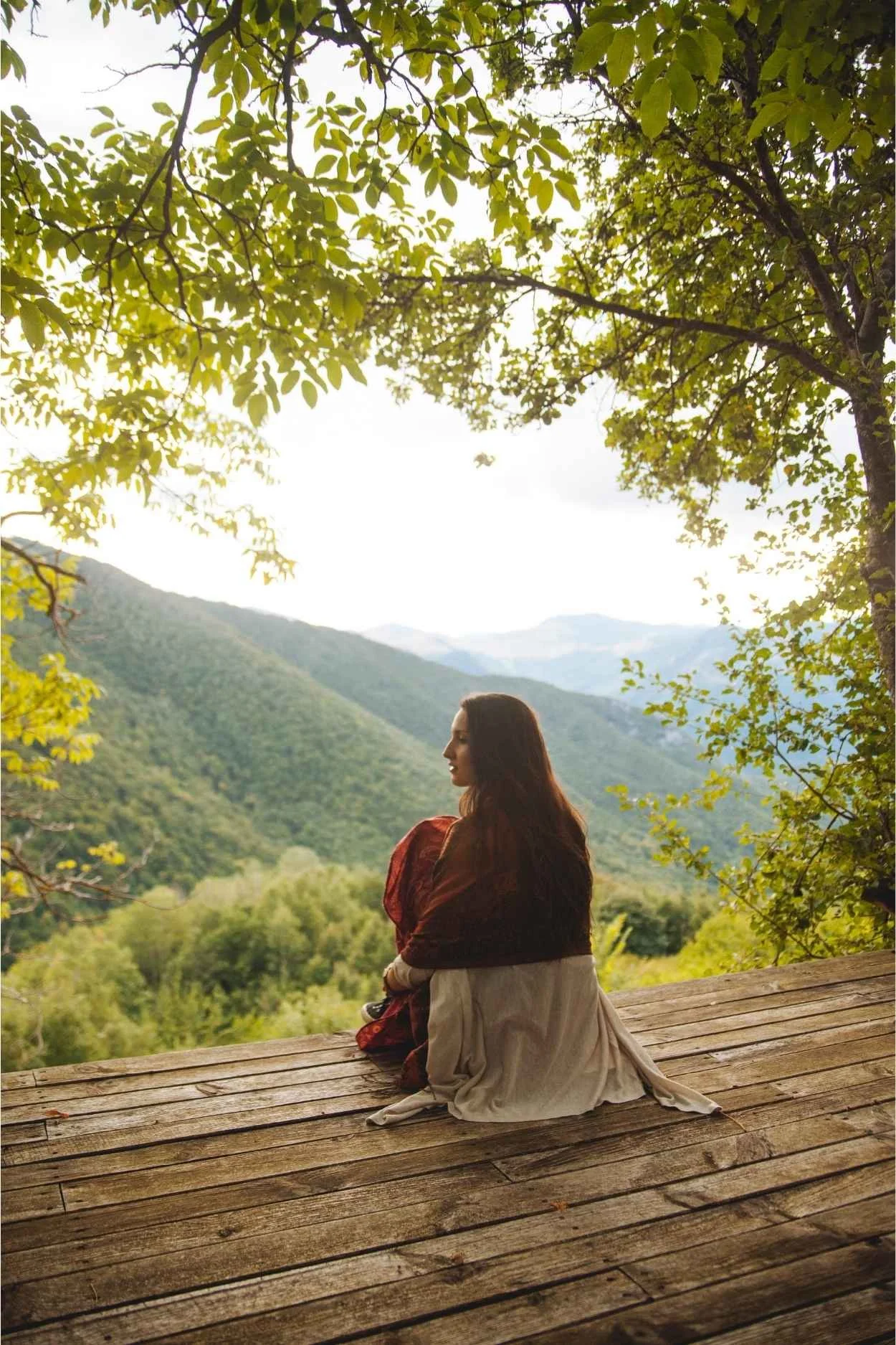 A woman sitting on a wooden deck overlooking a lush, green valley and mountains under a leaf-covered tree canopy.