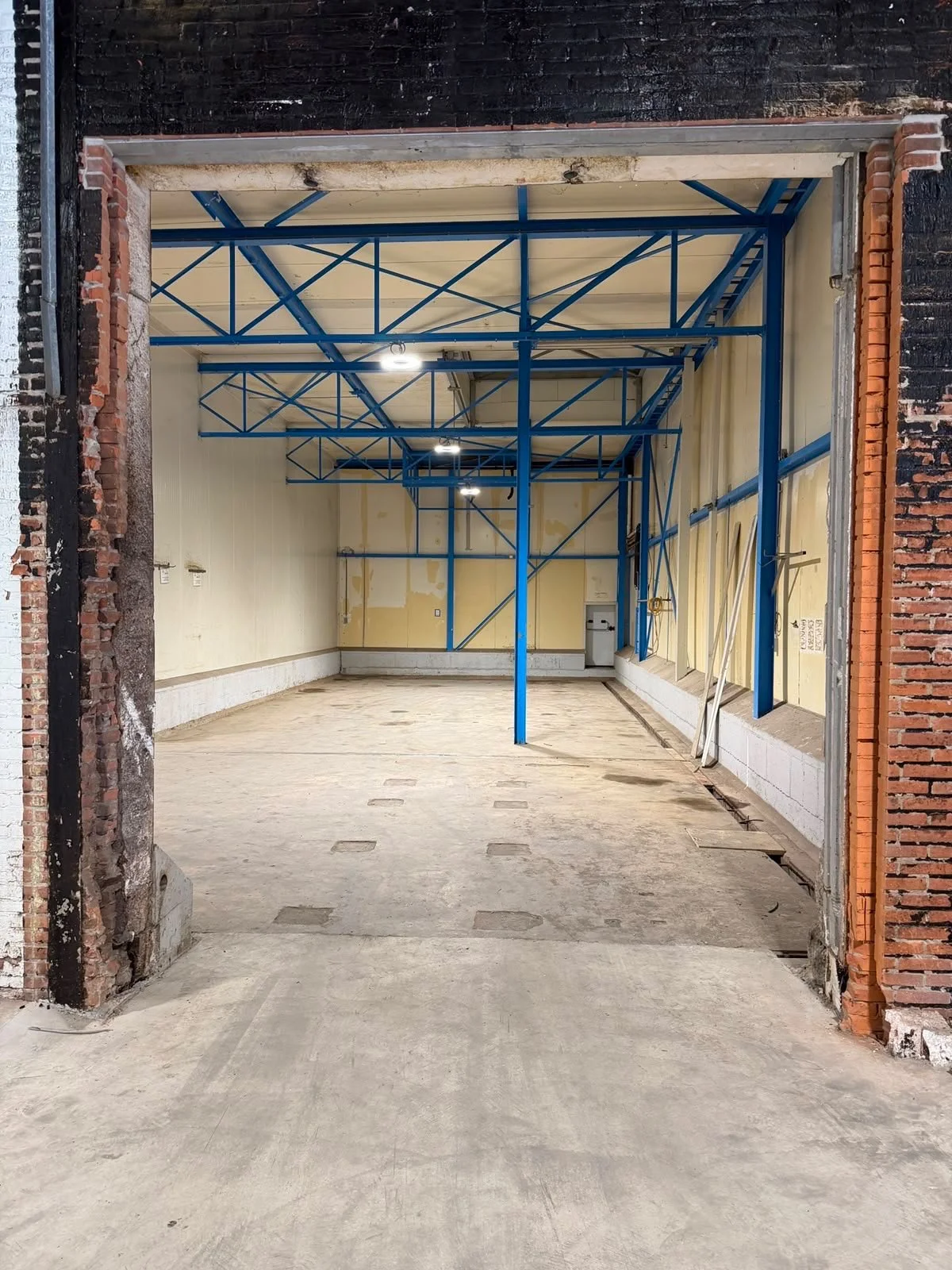 View of an indoor construction site with blue metal framing, unfinished walls, and exposed concrete floor, seen through an open doorway.