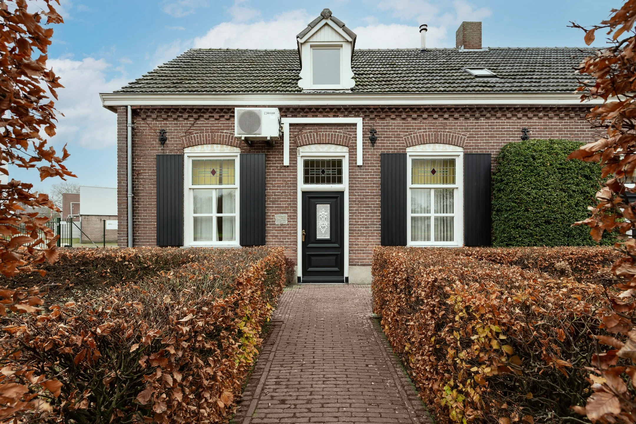 Front view of a brick house with black shutters, a pedestrian brick pathway, detached from the street, surrounded by a hedge and trees with brown autumn leaves.